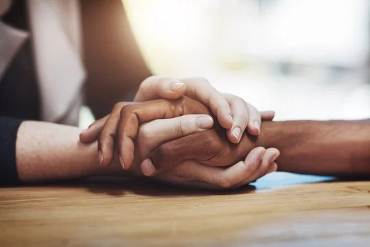 Close-up of two people holding hands on a wooden table, one with a lighter skin tone and the other with a darker skin tone, in a comforting gesture.