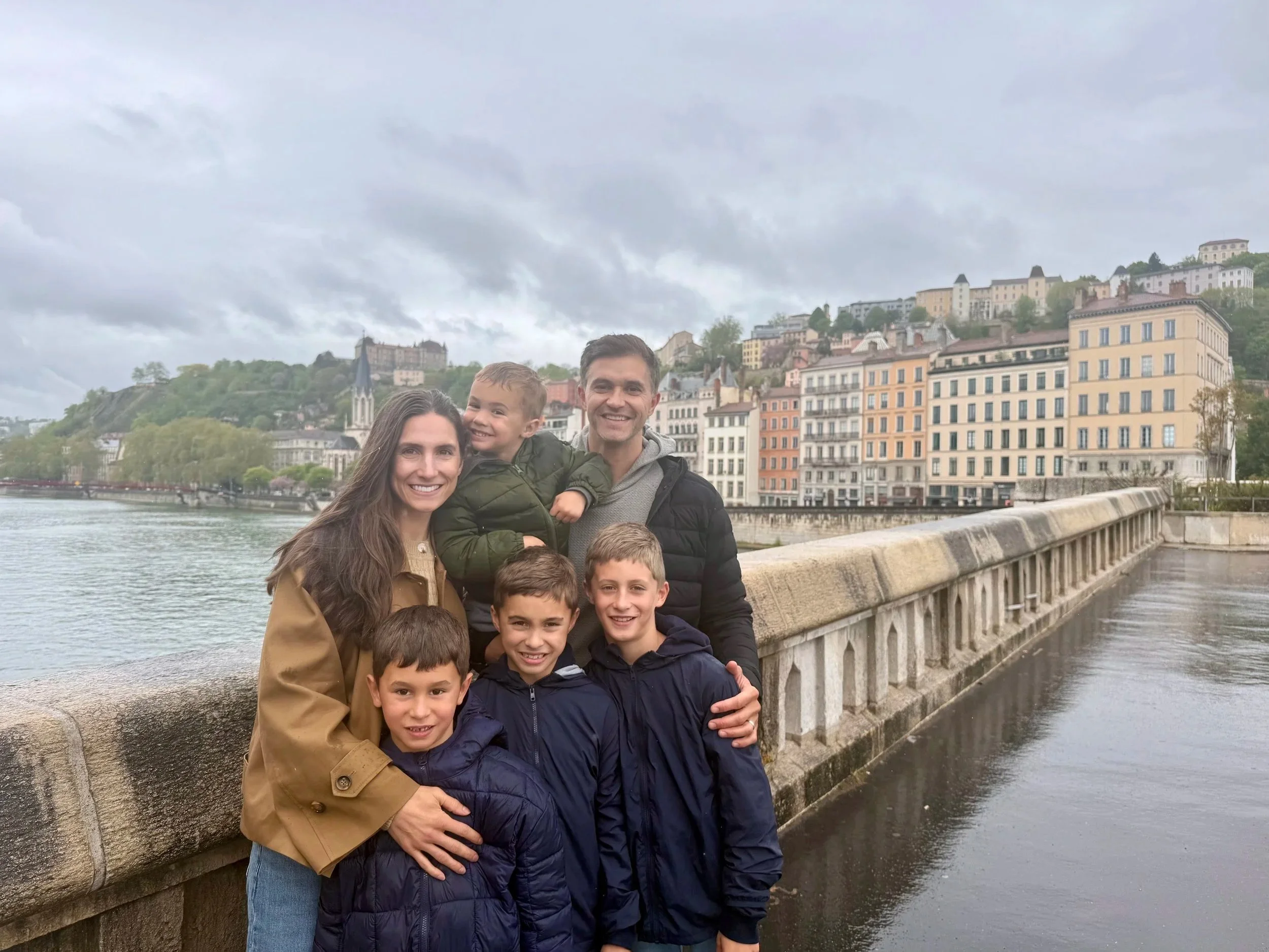 Trent Meacham and his family of six posing on a bridge in Lyon, France during a recent return to Europe with colorful buildings and hills in the background.