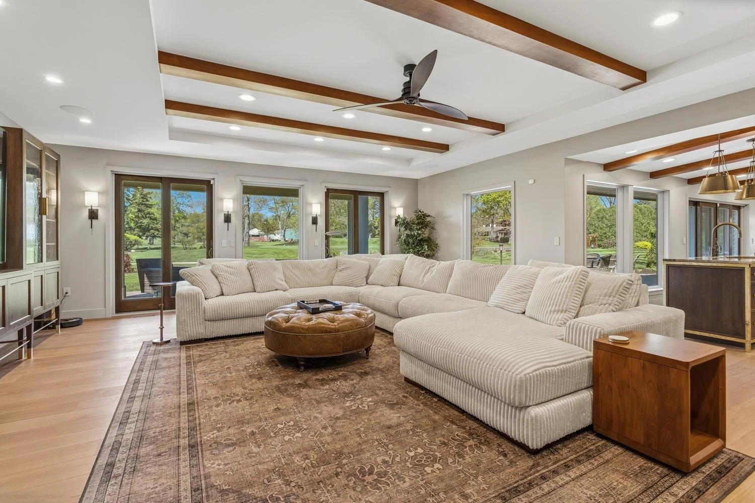 Realtor Trent Meacham's luxury house listing of living room with a large beige sectional sofa, a round leather ottoman, a brown side table, large windows showing a yard, and a ceiling with wooden beams.