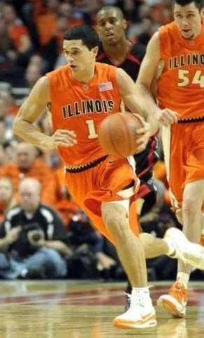 Real Estate agent Trent Meacham in an Illinois team uniform dribbling the basketball during a game, with two other players nearby.