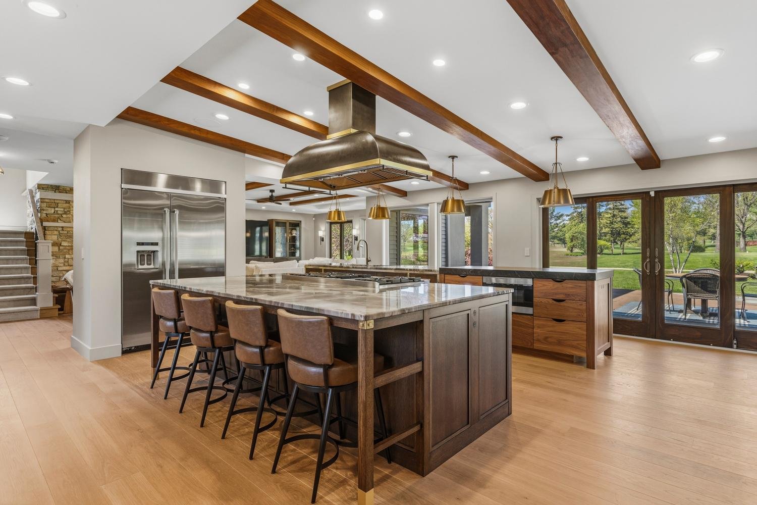 Trent Meacham luxury house listing in Champaign-Urbana of modern kitchen with wooden accents, stainless steel appliances, a large island with barstools, and sliding glass doors leading to an outdoor patio view of a green yard.