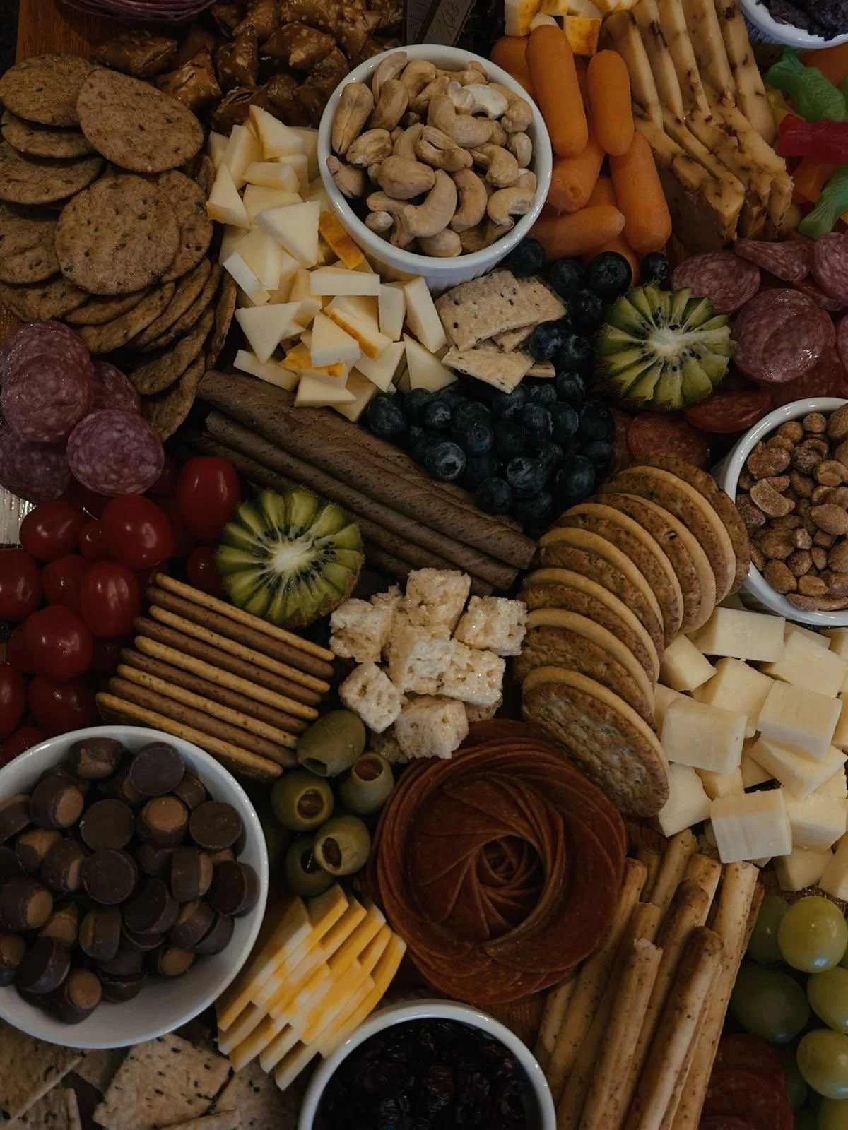 Assorted cheese cubes, grapes, cherry tomatoes, crackers, nuts, salami slices, chocolate candies, green olives, and a small container of jam on a charcuterie board.