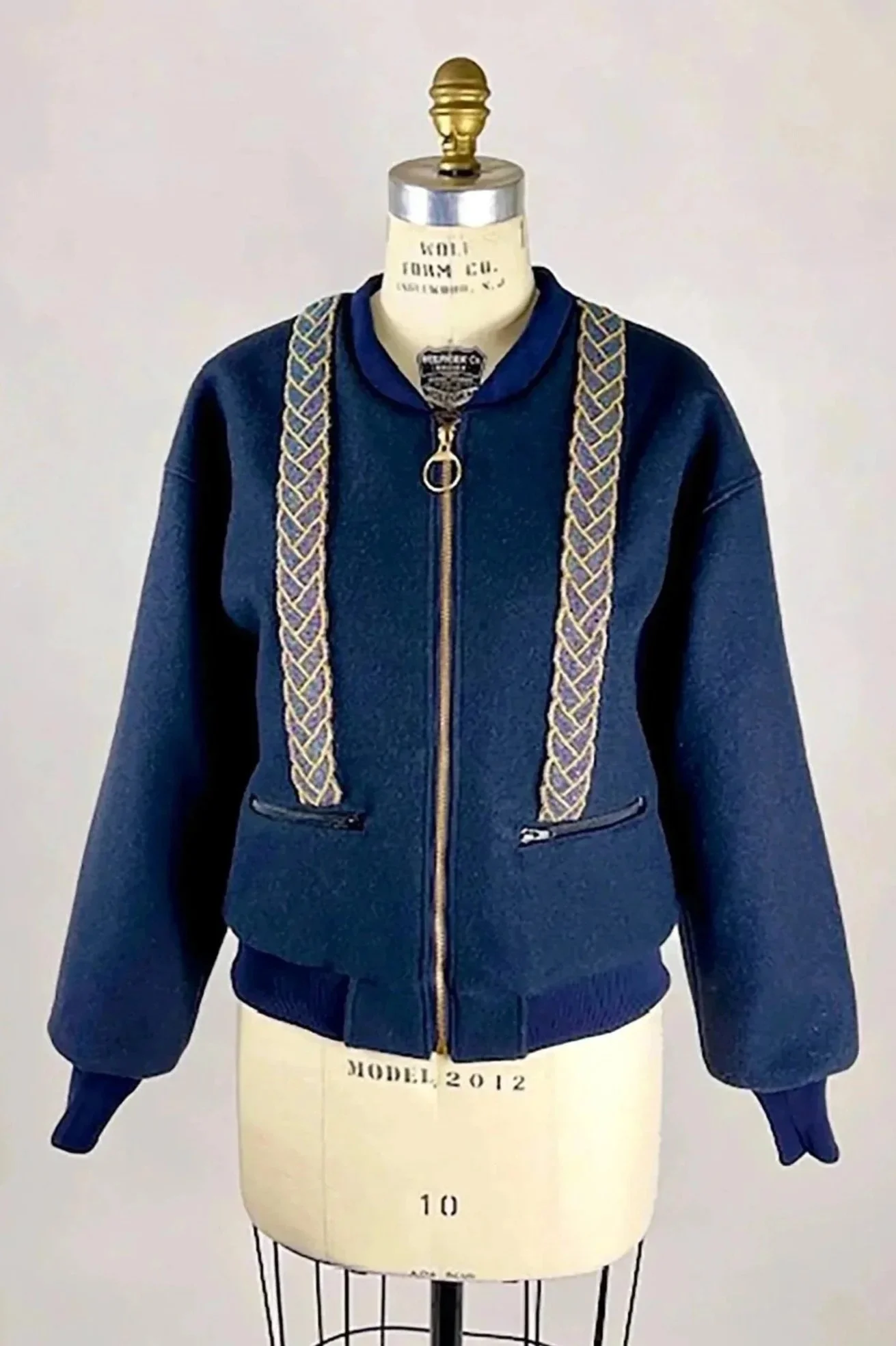 A mannequin wearing a dark blue zip-up jacket with gold braided straps draped over the shoulders, displayed on a stand marked 'MODEL 2012'.