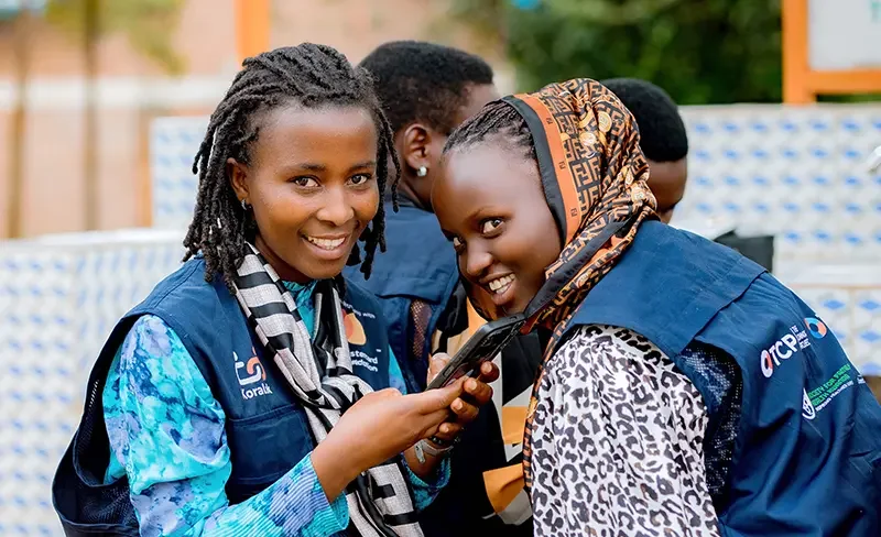 2 young women sitting next to each other smiling wearing KoraLink agent vests and holding a cell phone