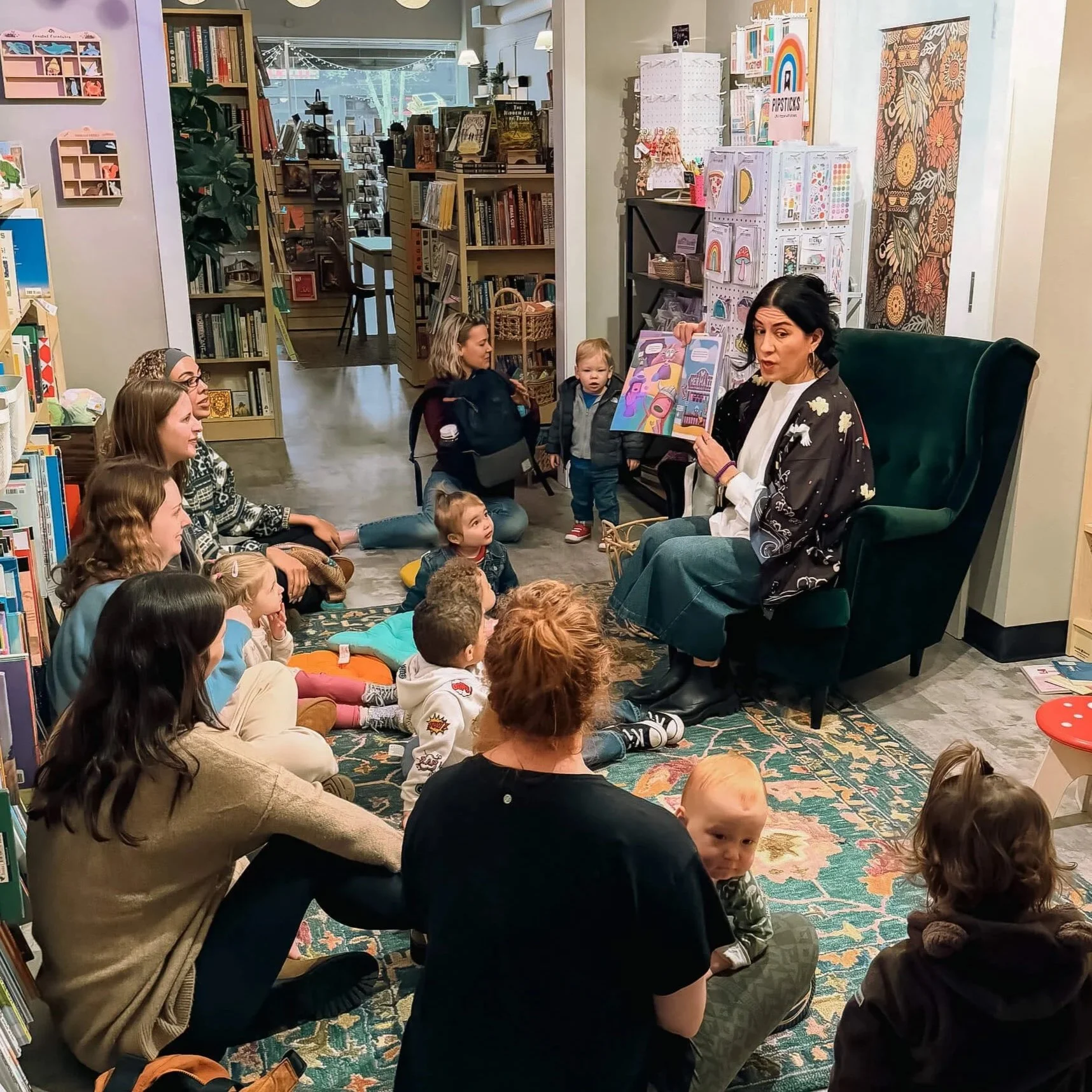 Toddler story time event at Paper Boat Booksellers in West Seattle, with young children and caregivers gathered on a rug while a reader holds up a picture book in the colorful children's section