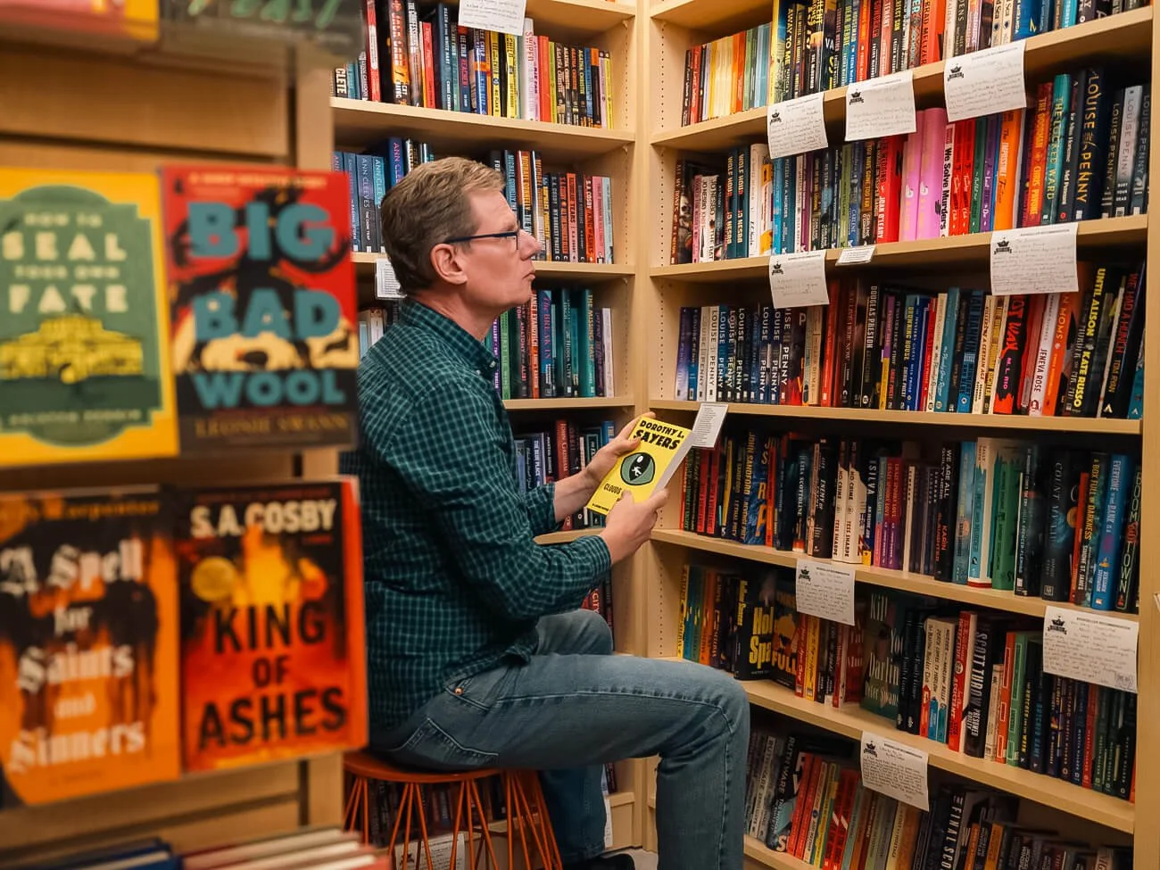 A customer seated on a stool reading a Dorothy L. Sayers novel in the mystery section of Paper Boat Booksellers, surrounded by shelves of crime and thriller books