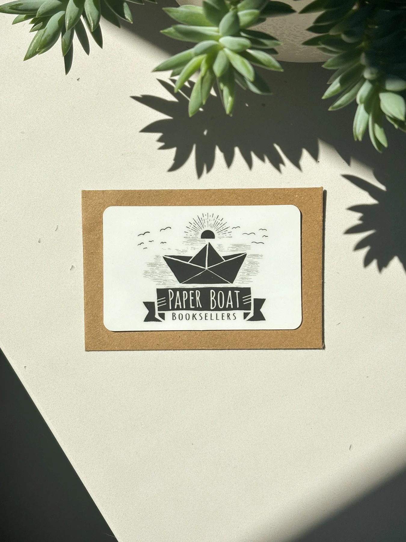 Paper Boat Booksellers gift card next to a succulent plant making interesting shadows on the counter