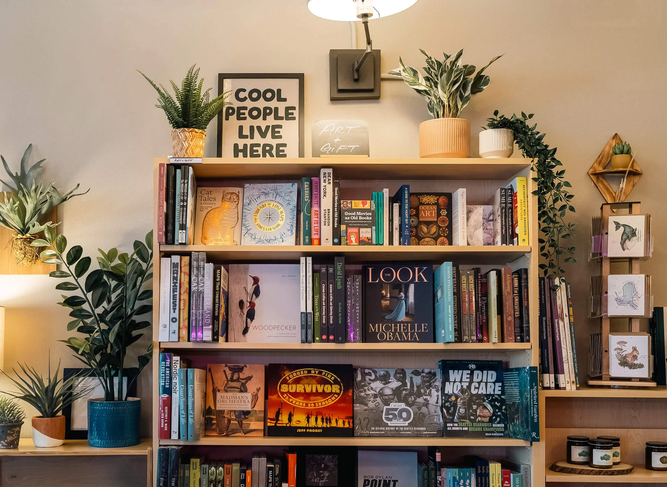 Art and gift section at Paper Boat Booksellers decorated with lush houseplants, featuring coffee table books including Michelle Obama's The Look, Woodpecker, and Survivor, alongside a framed "Cool People Live Here" print