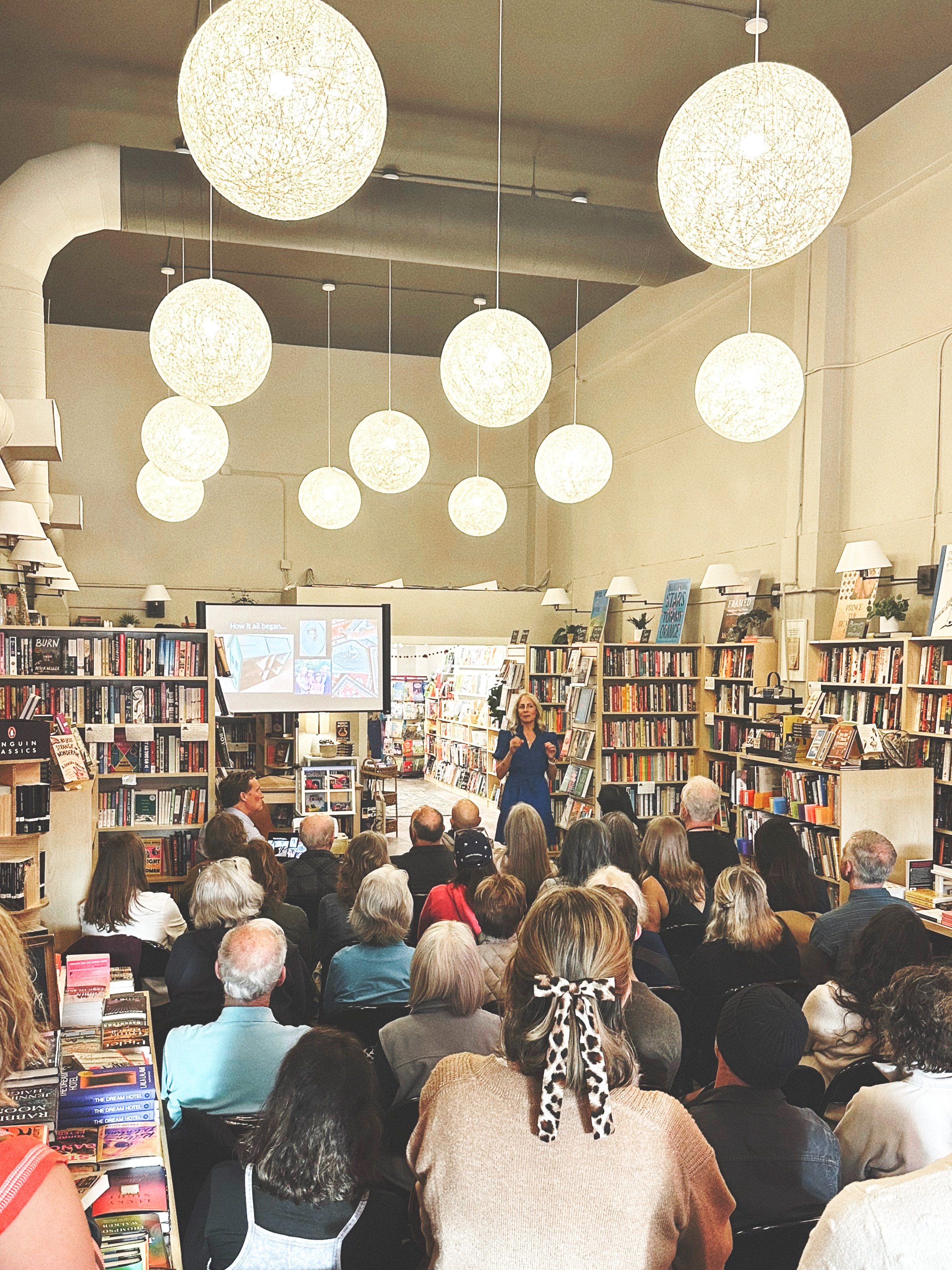 Packed author talk at Paper Boat Booksellers in West Seattle, with a crowd seated facing a speaker presenting in front of a projection screen, surrounded by bookshelves and globe pendant lights