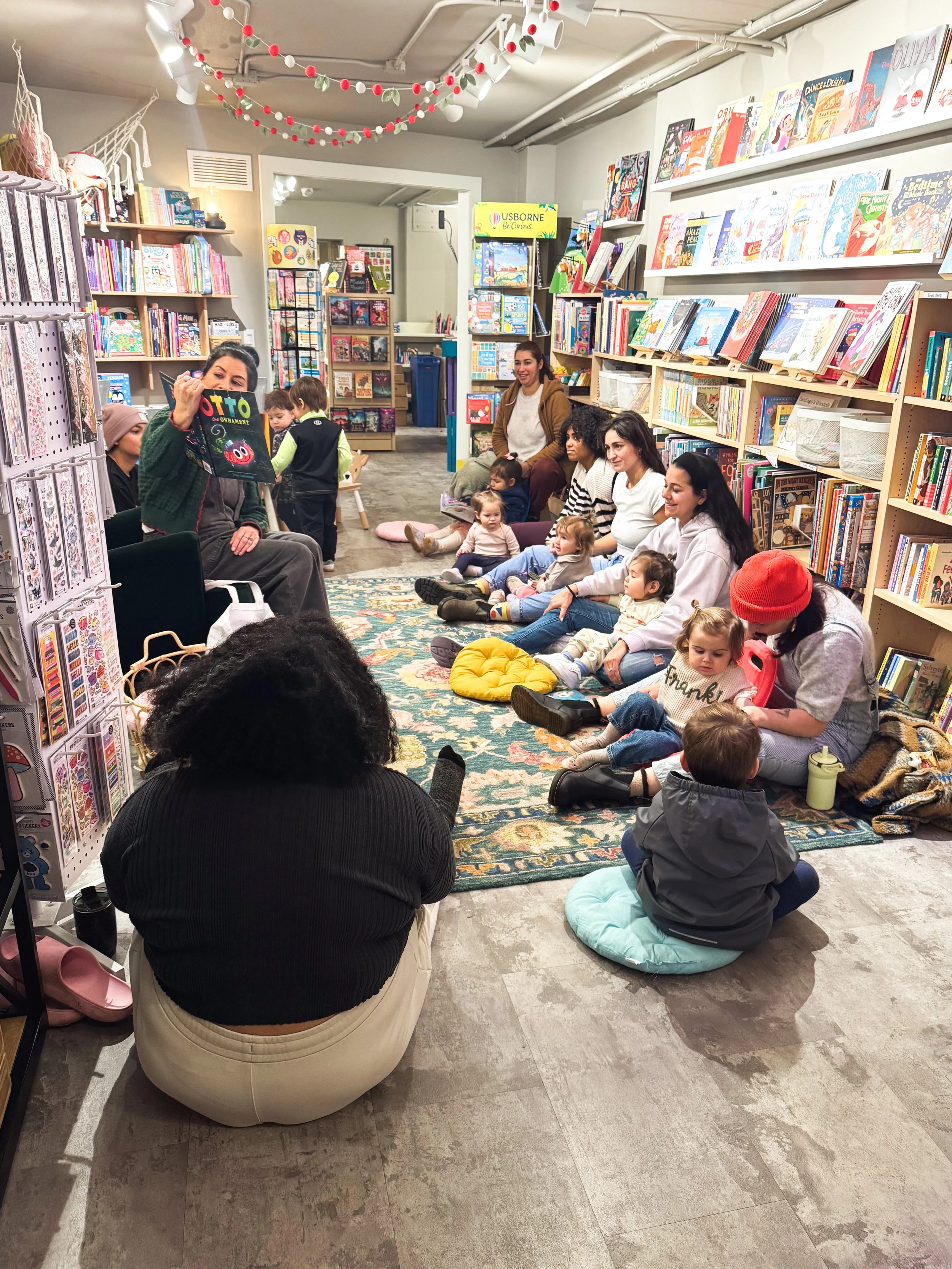 Toddler story time event at Paper Boat Booksellers in West Seattle, with young children and caregivers gathered on a rug while a reader holds up a picture book in the colorful children's section