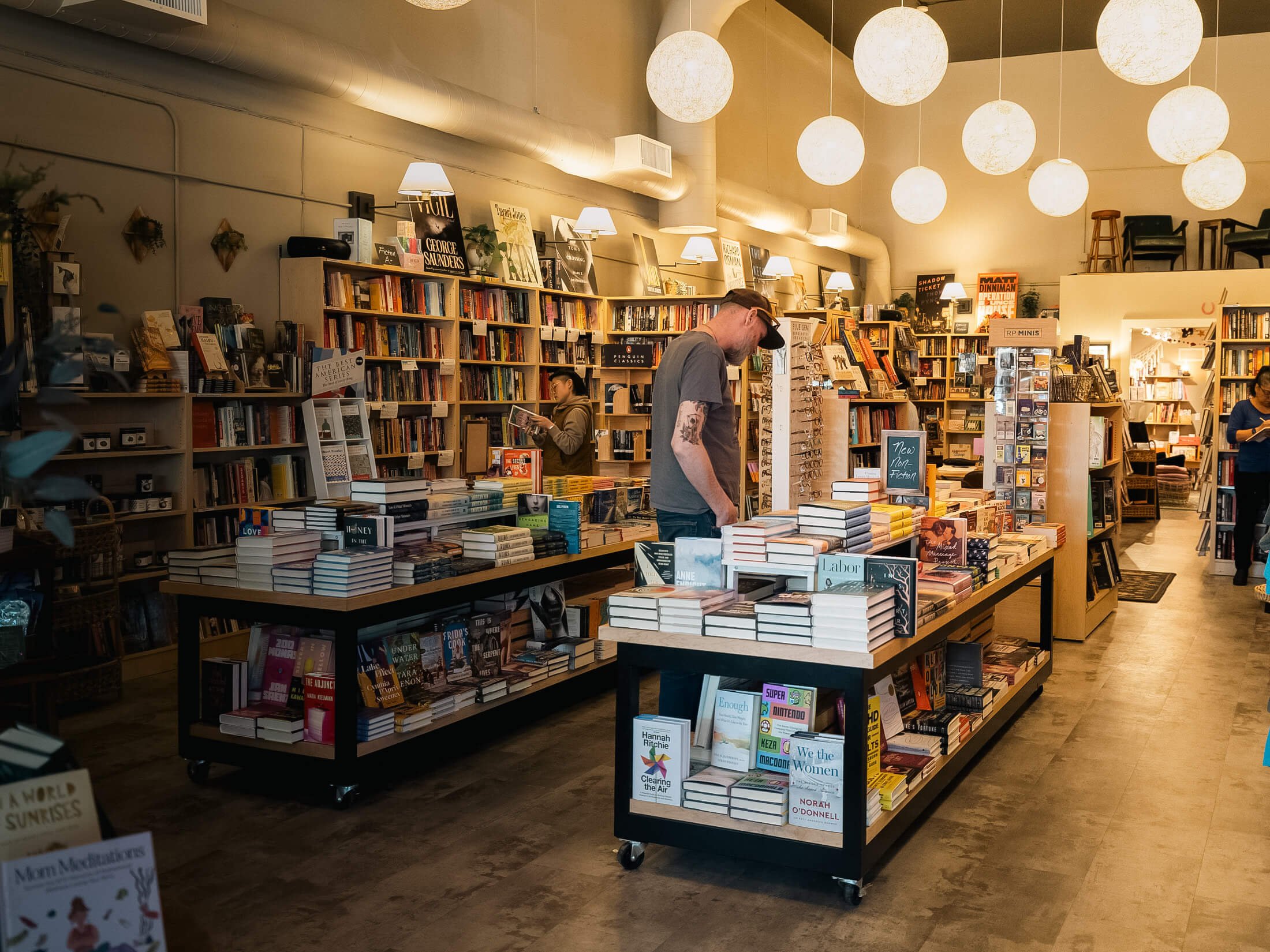 Wide interior view of Paper Boat Booksellers in West Seattle with customers browsing table displays and shelves under warm globe pendant lights