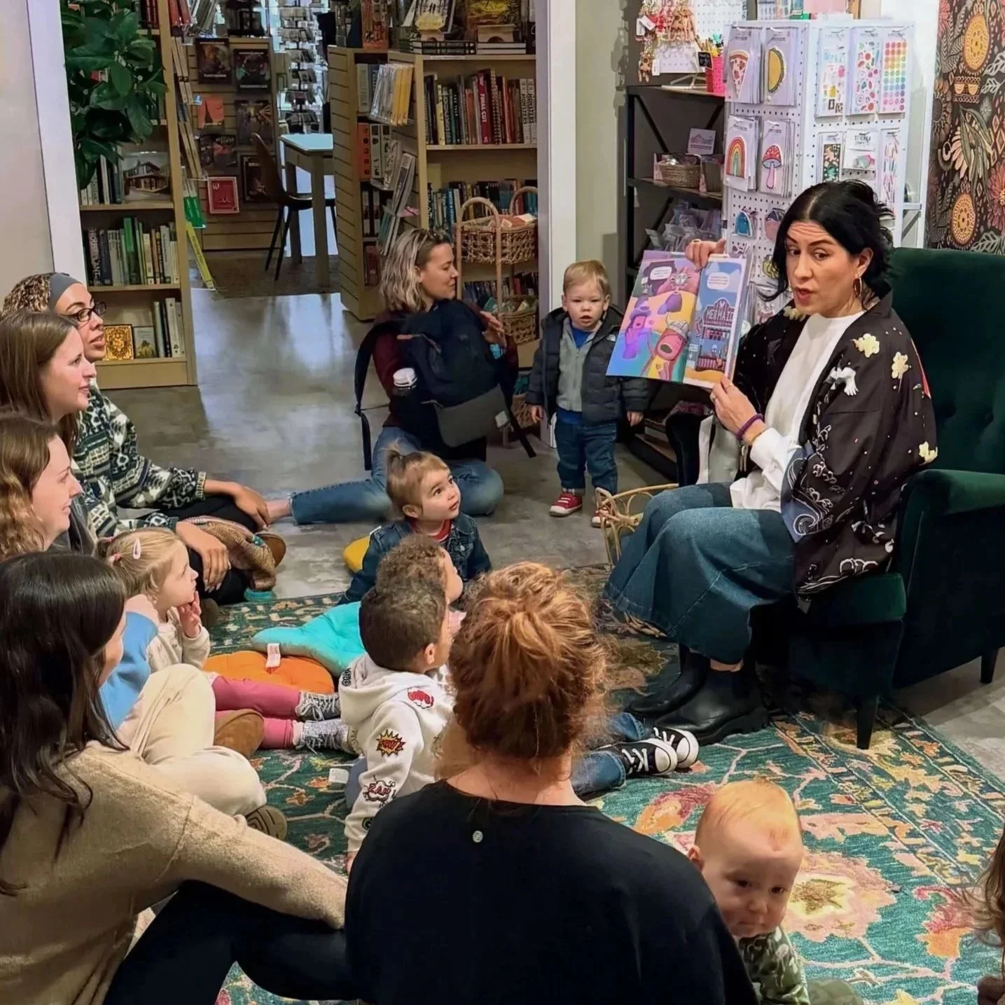 Toddler story time event at Paper Boat Booksellers in West Seattle, with young children and caregivers gathered on a rug while a reader holds up a picture book in the colorful children's section