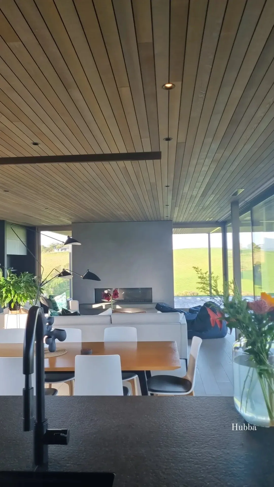 Modern living room with wooden ceiling, large windows showing green landscape, white furniture, potted plants, and a fireplace.