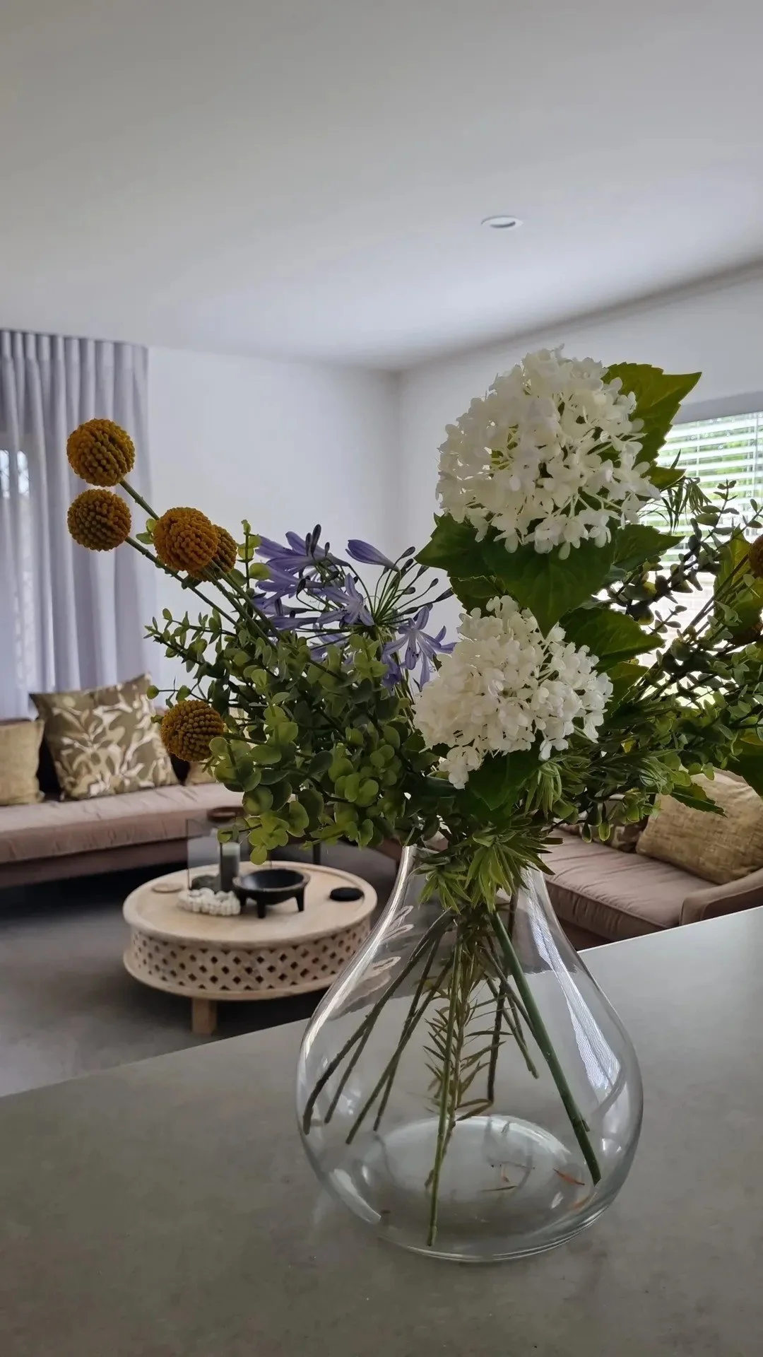 A clear glass vase filled with white and purple flowers and green foliage, placed on a table in a bright living room.