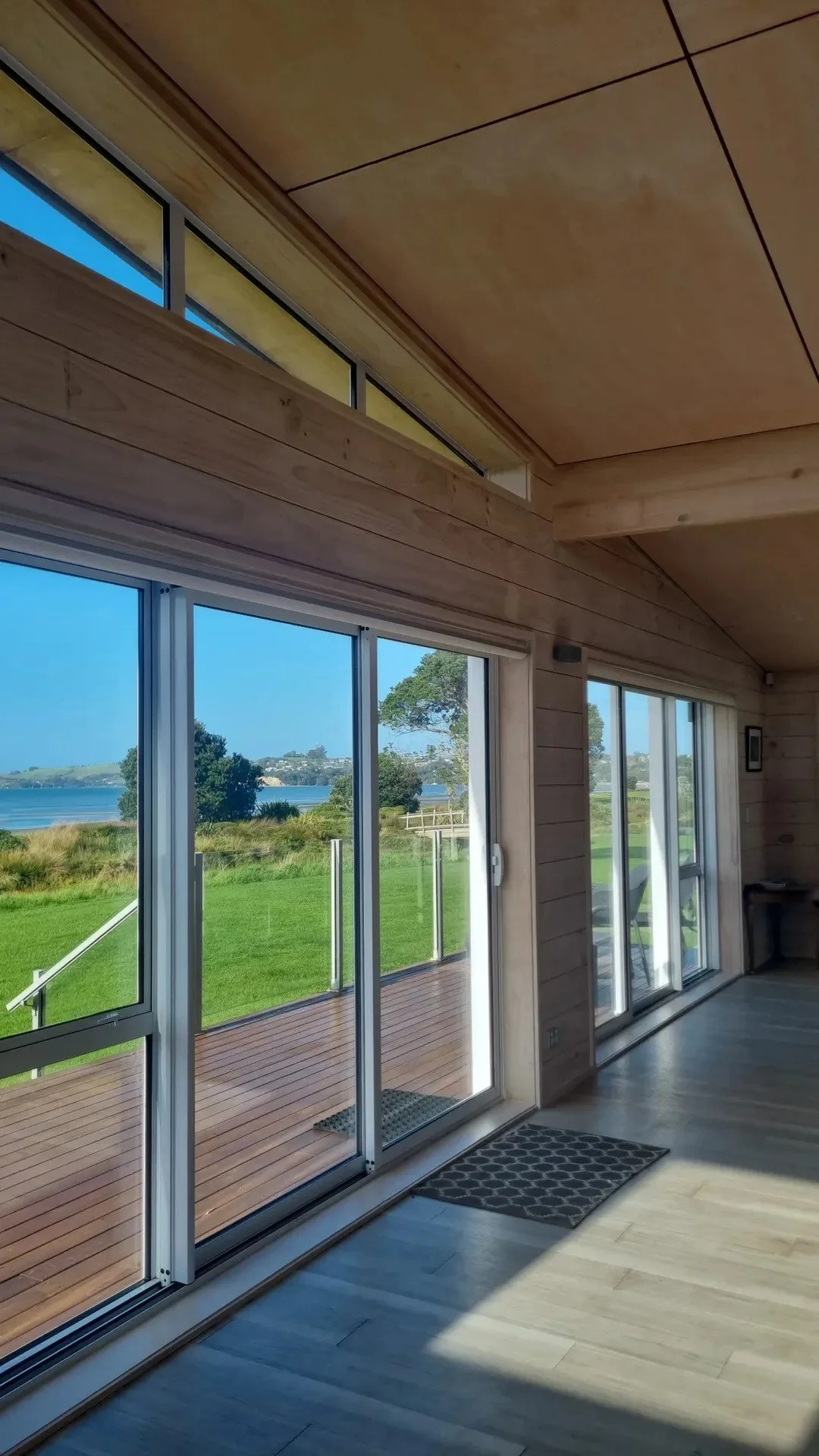 Interior view of a room with large glass sliding doors and windows, revealing a lush green outdoor landscape with a body of water and trees in the distance, and a small wooden deck outside.