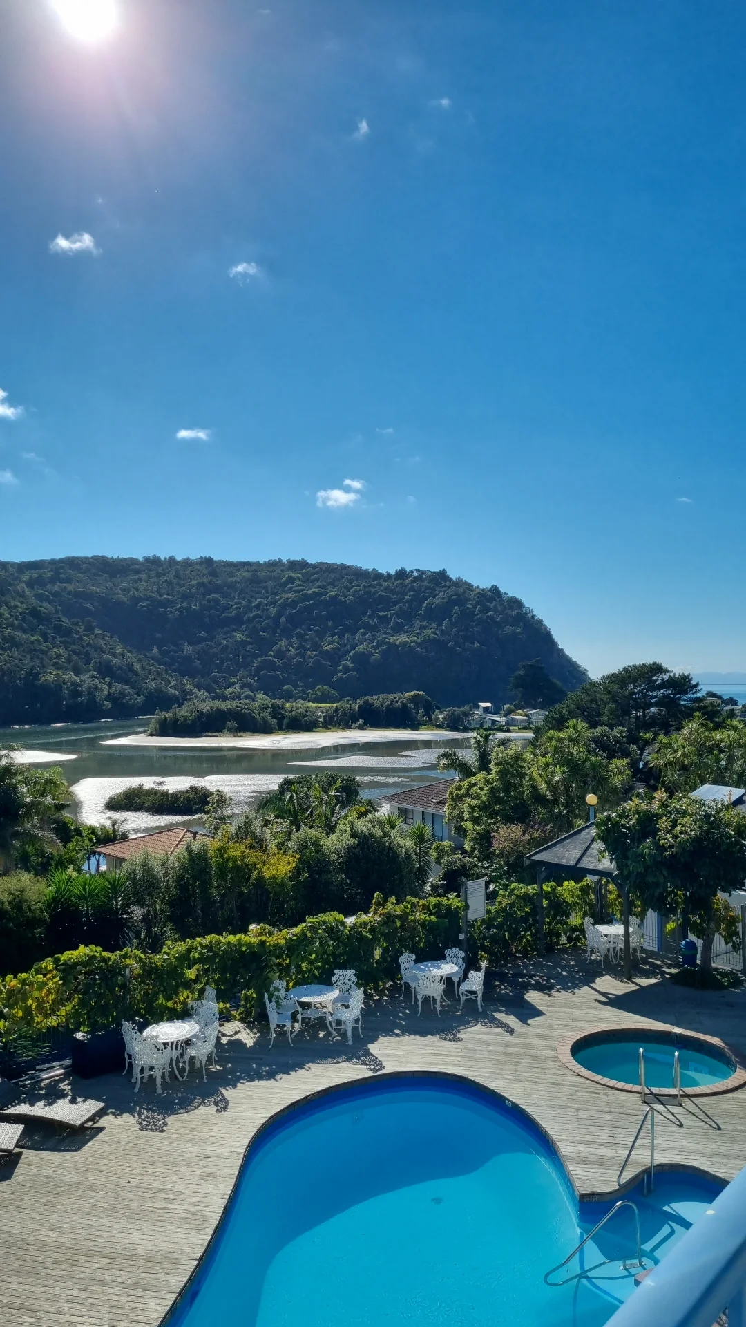 A scenic outdoor view of a hillside overlooking a river, with houses and lush greenery, under a clear blue sky with a few scattered clouds and the sun shining.