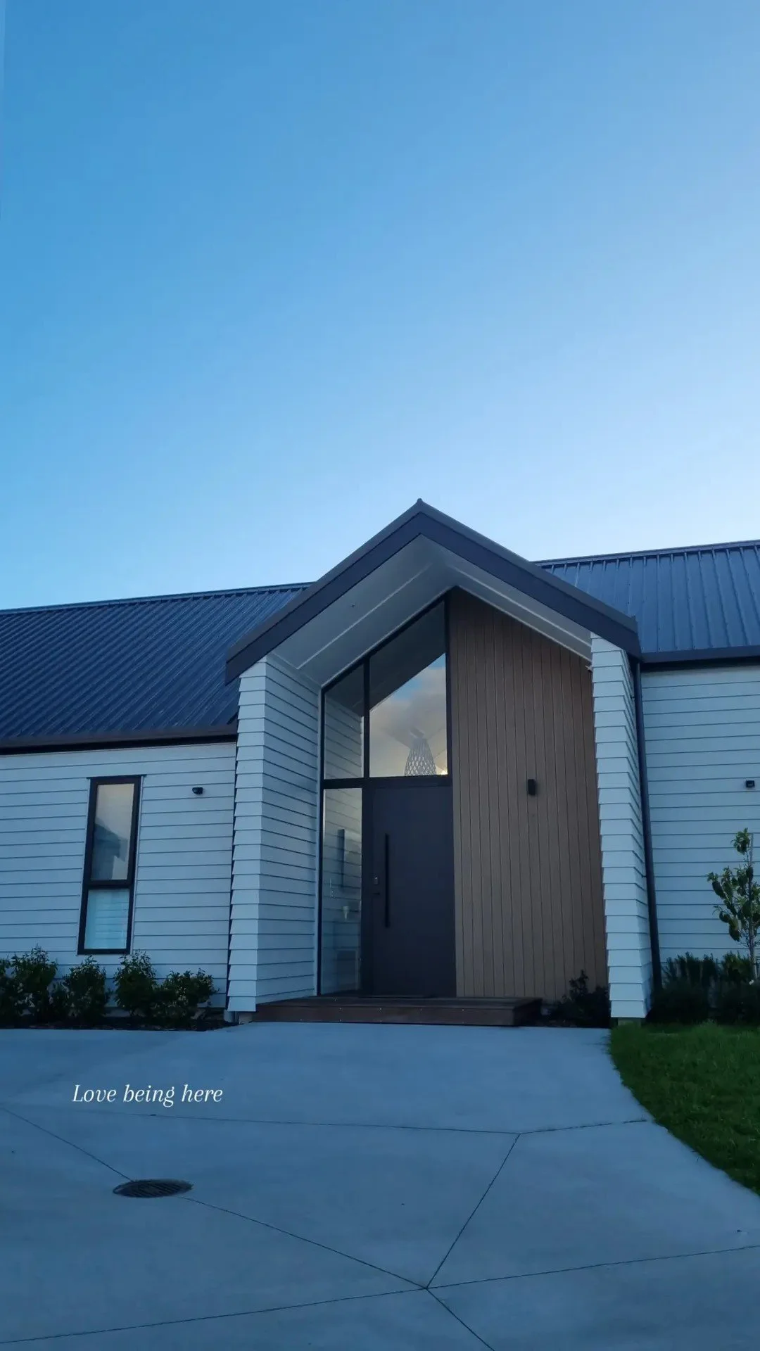 Modern house with a sloped roof, large glass windows, and a minimalist entrance. The sky is clear and blue, with some plants and concrete pavement in front. Text at the bottom left reads "Love being here."