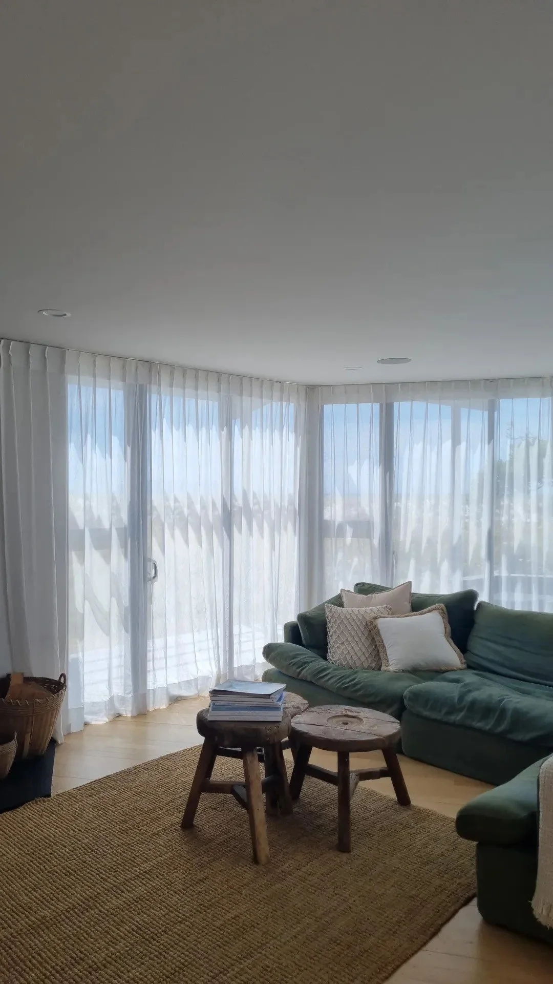 Living room with sheer white curtains, green sofa with pillows, small wooden side tables, and a woven rug.