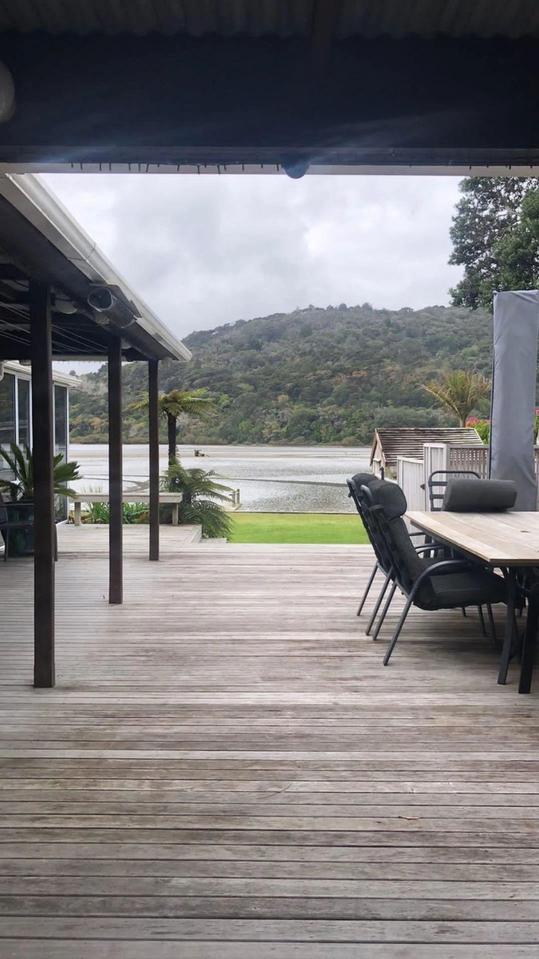 A wooden deck overlooking a lake with a house in the background, trees, and hills under a cloudy sky.