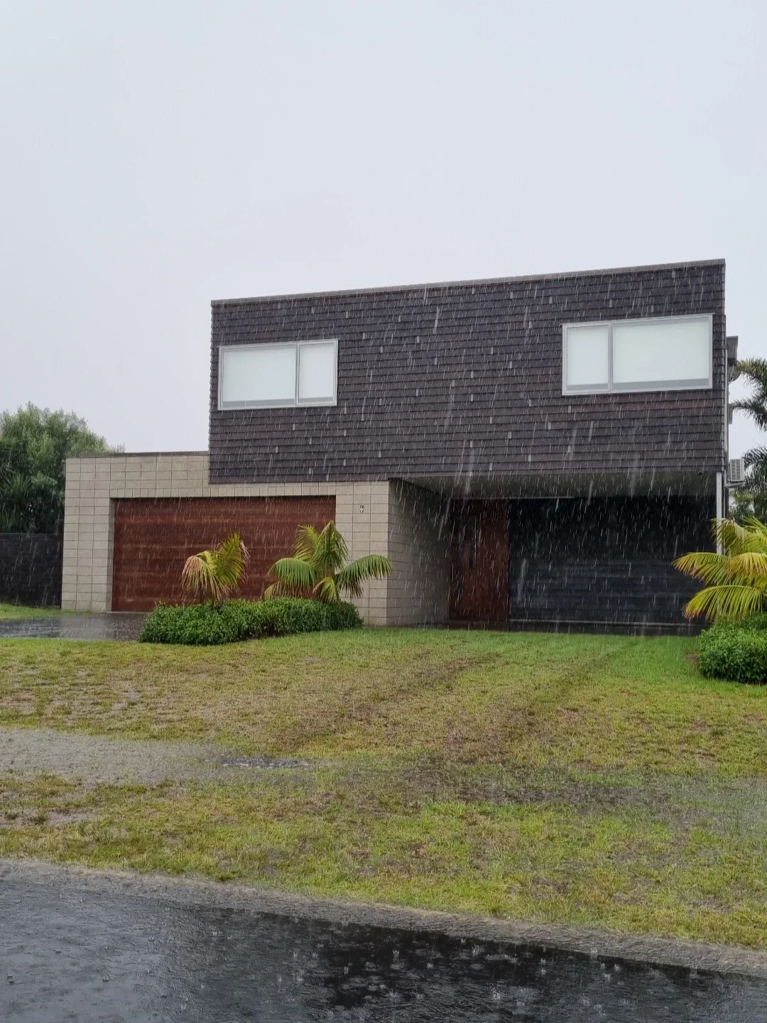 Modern house with brown and black garage doors, gray upper level with large windows, green lawn, and rain falling.