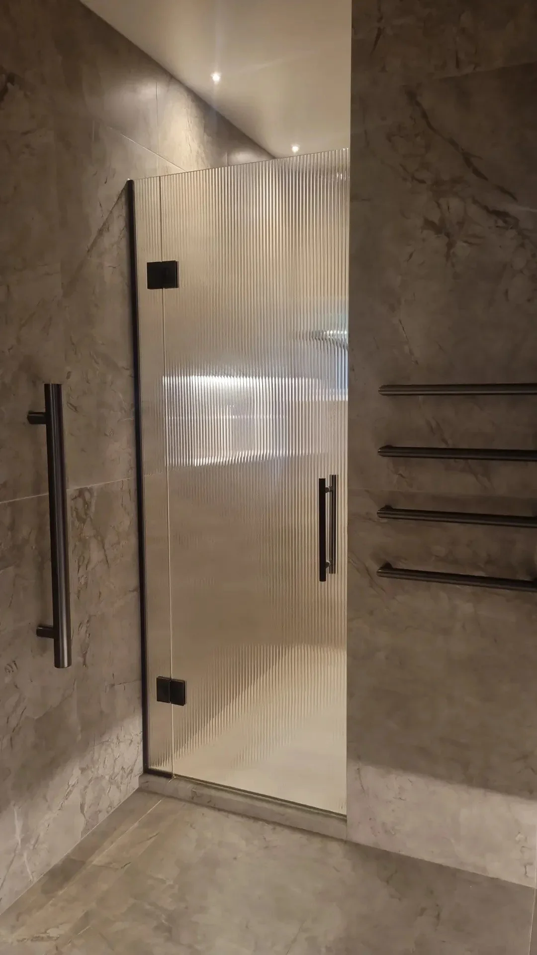 A restroom stall with a faceted metal door, two small black hinges, a black handle, and marble walls and floor.