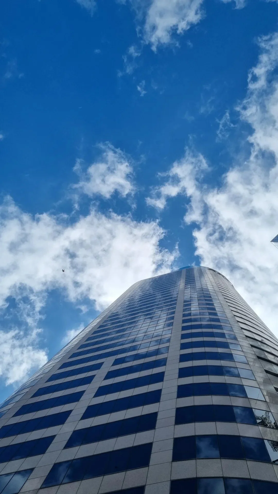A tall modern skyscraper with a curved glass facade reflecting the sky, taken from ground level looking up, with a blue sky and scattered clouds in the background.