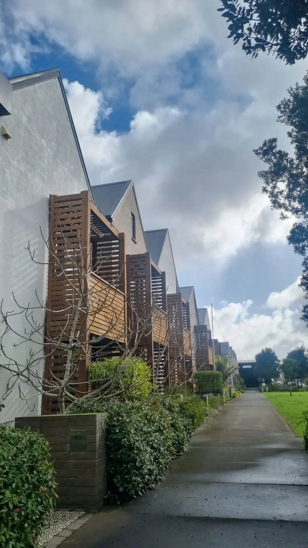 Row of modern townhouses with wooden balconies and leafless trees in front, under a partly cloudy sky.