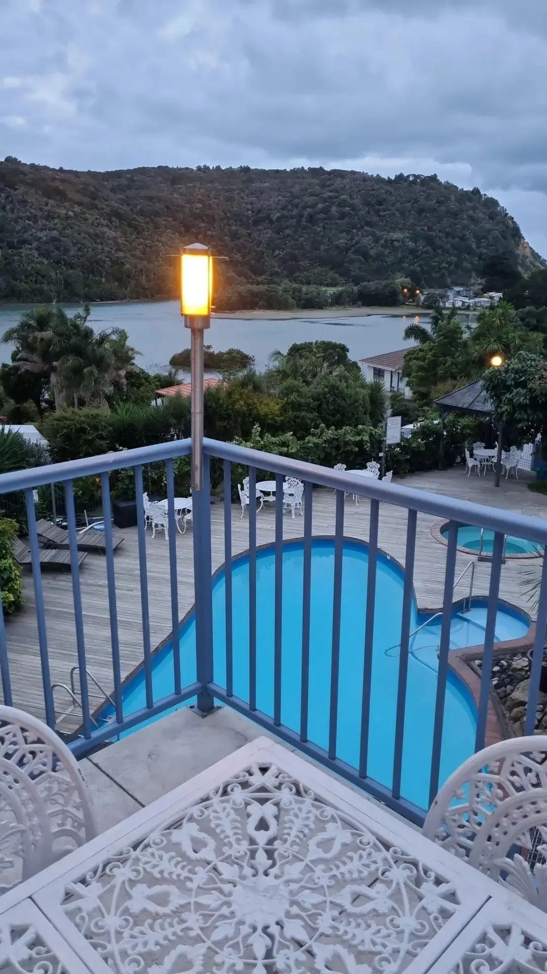 View from a balcony showing a swimming pool, white garden furniture, trees, a river, hills in the distance, and a cloudy sky.