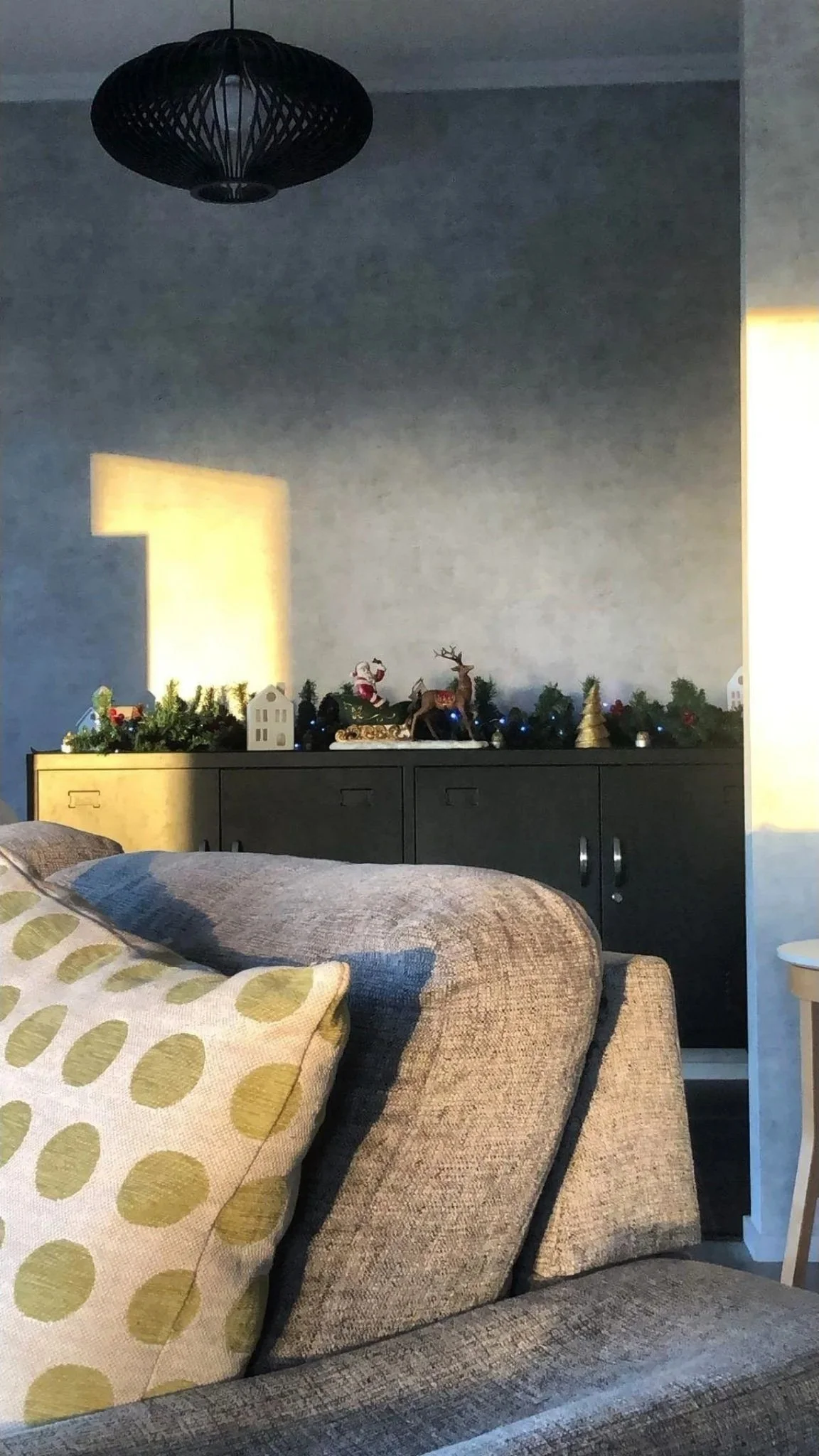Living room with beige fabric couch and cushions, black sideboard with Christmas decorations including a reindeer figure, Santa figure, small house, and greenery with ornaments, illuminated by yellow wall lights, and a dark ceiling light fixture.