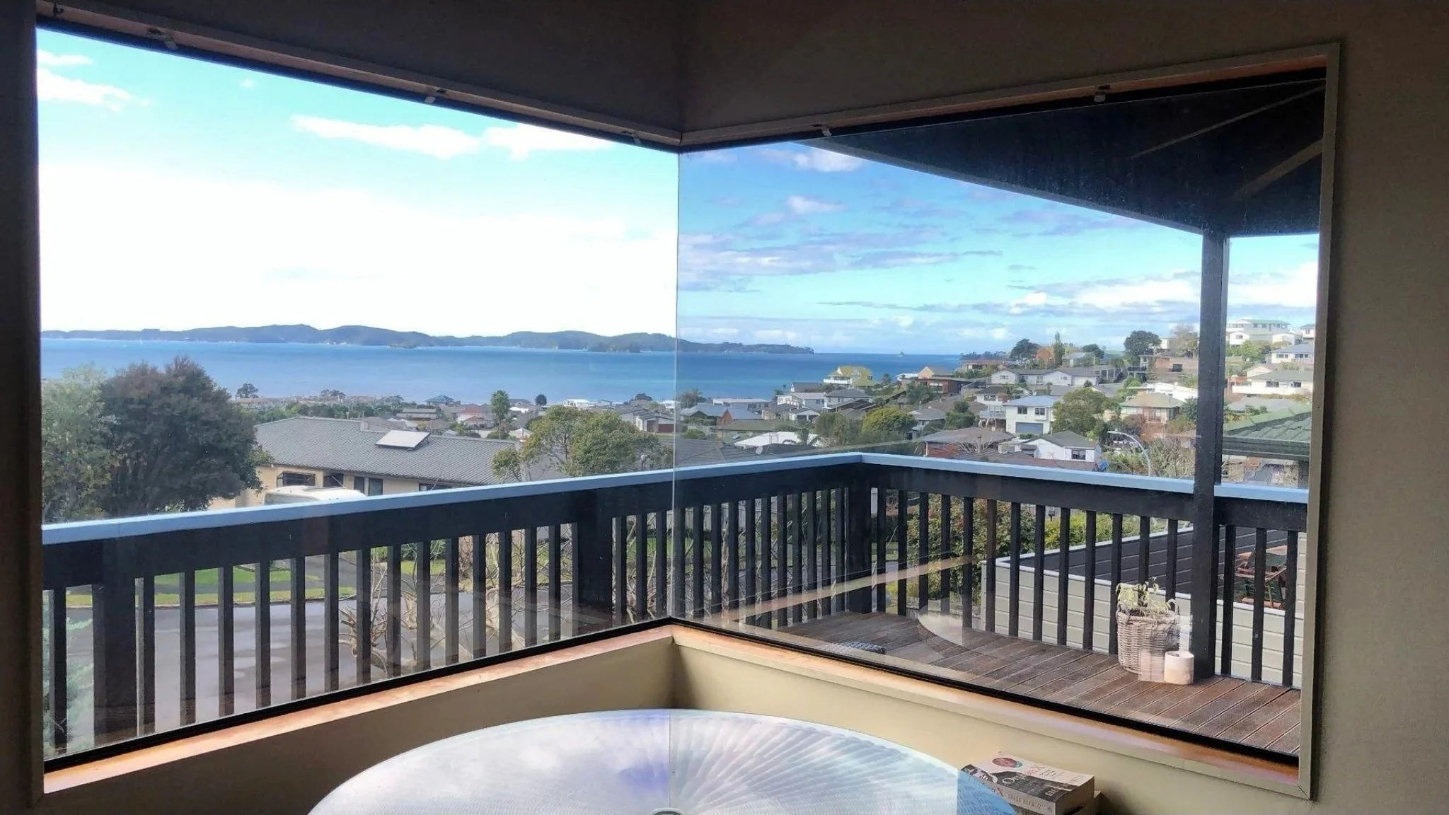 View from a balcony showing a neighborhood with houses, trees, and a body of water in the distance under a partly cloudy sky.