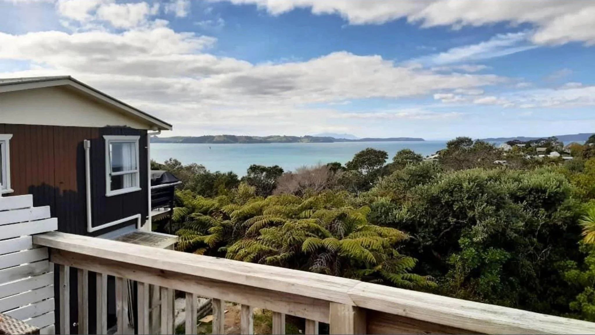 View of a coastal landscape with a house in the foreground, lush greenery, and a body of water with distant landforms under a partly cloudy sky.