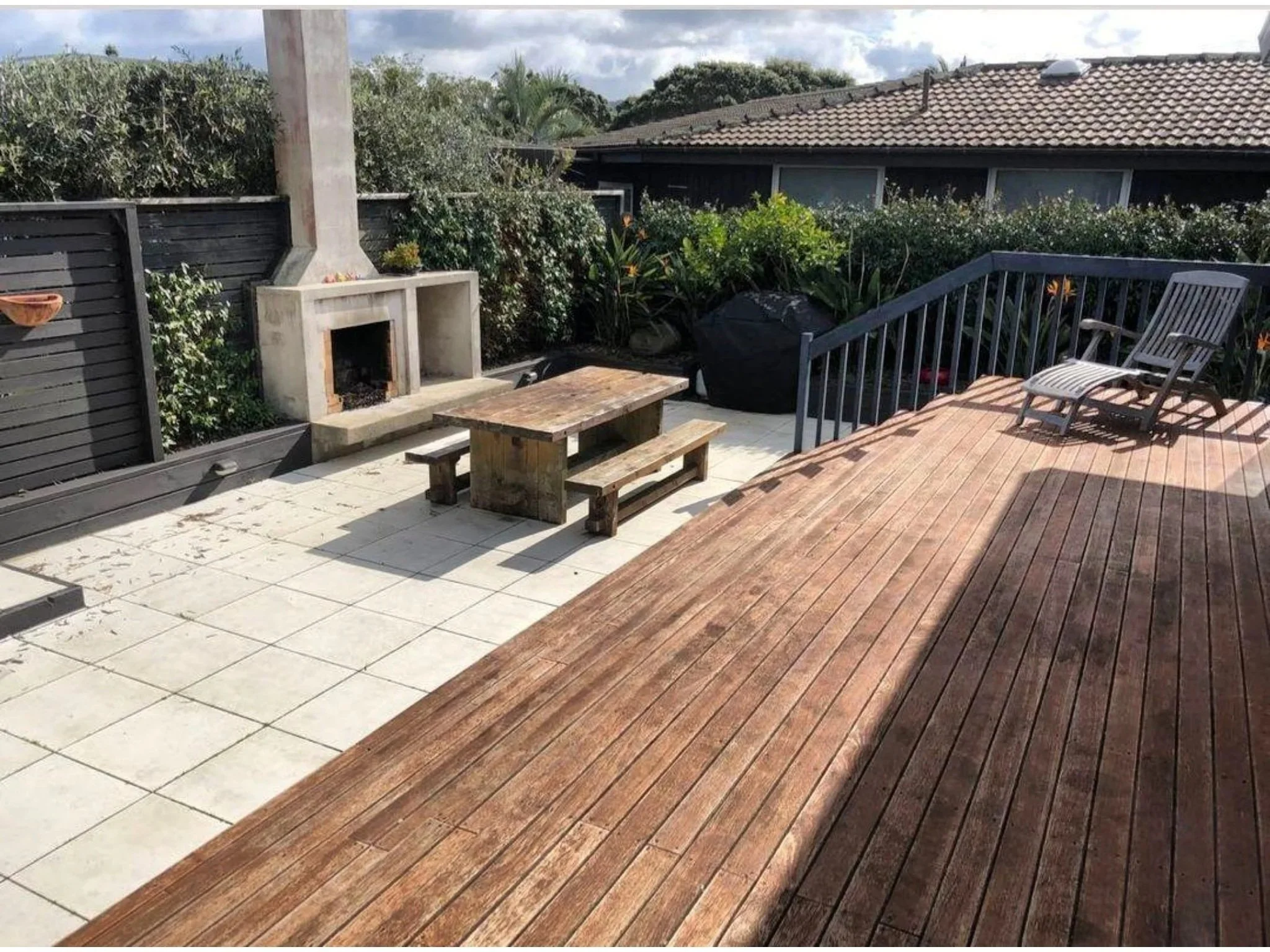 An outdoor patio area with a wooden deck, a small picnic table with benches, and a black chair. There is a concrete fireplace or grill with a chimney, and a black privacy fence. Surrounding the patio are green bushes and plants. A house with a tiled 