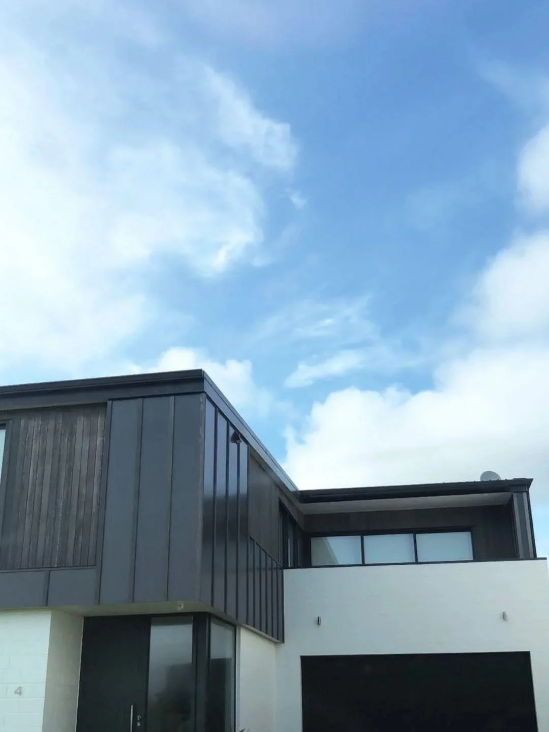 Modern building with dark gray metal panel exterior and large windows, against a sky with scattered clouds.
