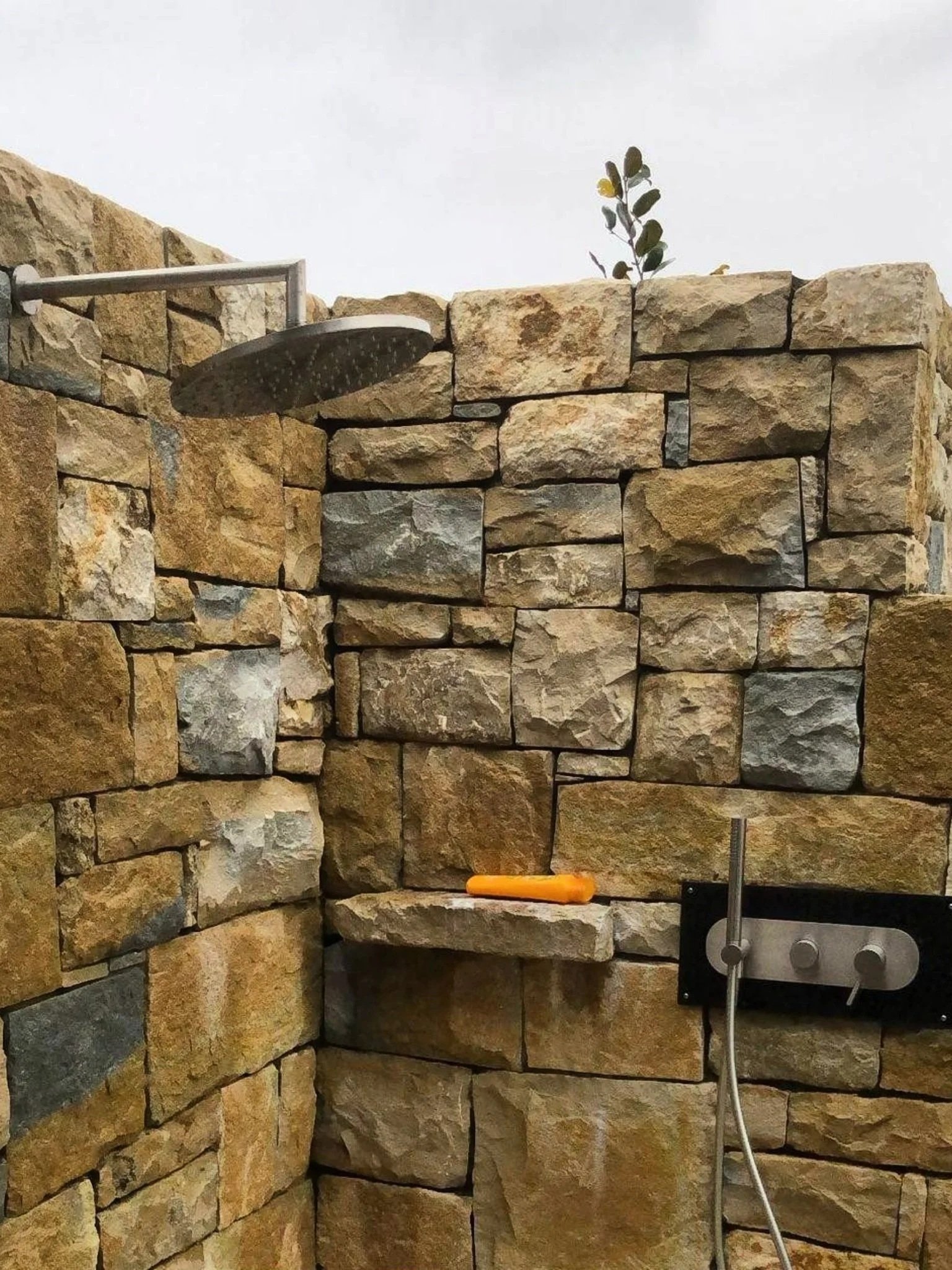 An outdoor shower with a stone wall background, a metal showerhead, and a control panel with a blue, orange, and gray handle.
