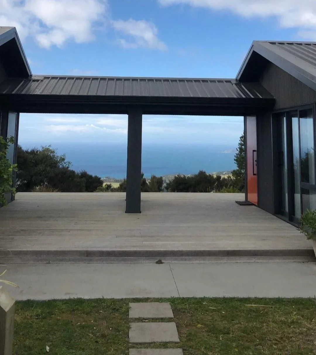 View through a modern patio with dark siding, showing a landscape with trees and the ocean in the distance, under a blue sky with clouds.