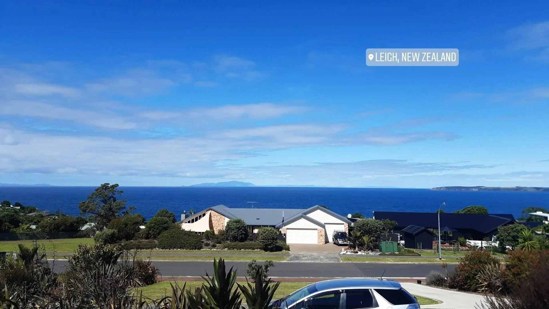 A residential neighborhood near the coast of Leigh, New Zealand, with houses, trees, and a view of the ocean and distant islands under a partly cloudy sky.
