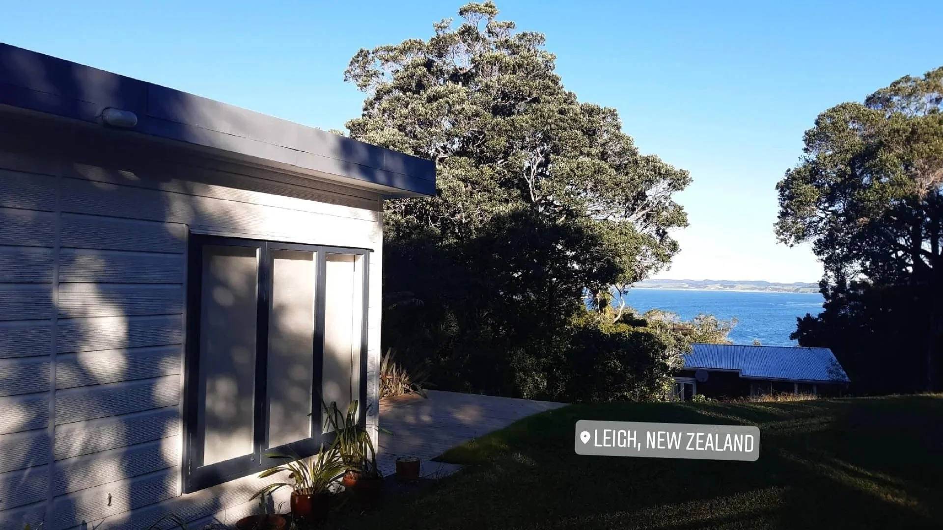 A house with grey siding and a white door, potted plants outside, trees in the background, overlooking a body of water in Leigh, New Zealand.