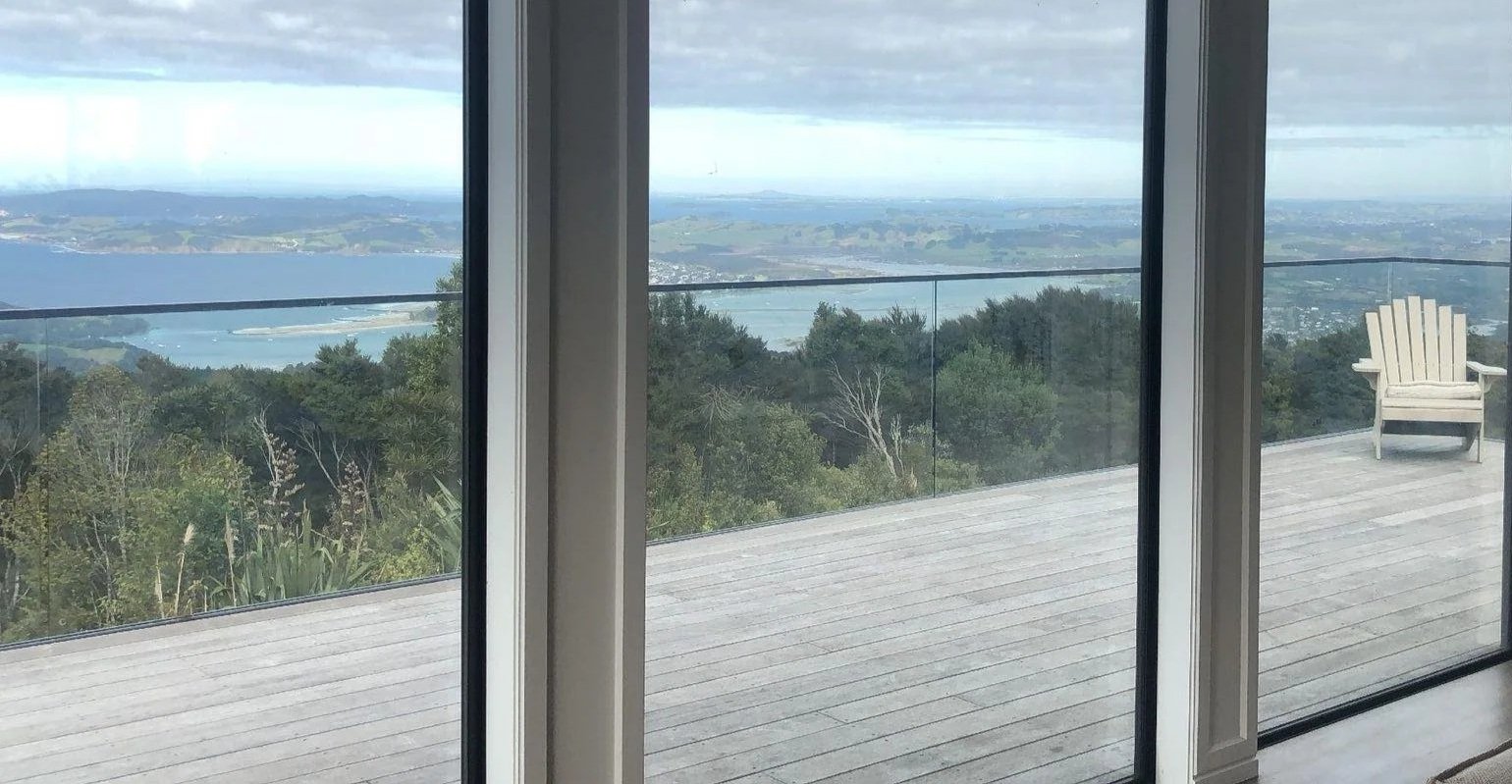 View of a body of water and distant hills seen through large sliding glass doors, with a wooden deck and a white chair outside.