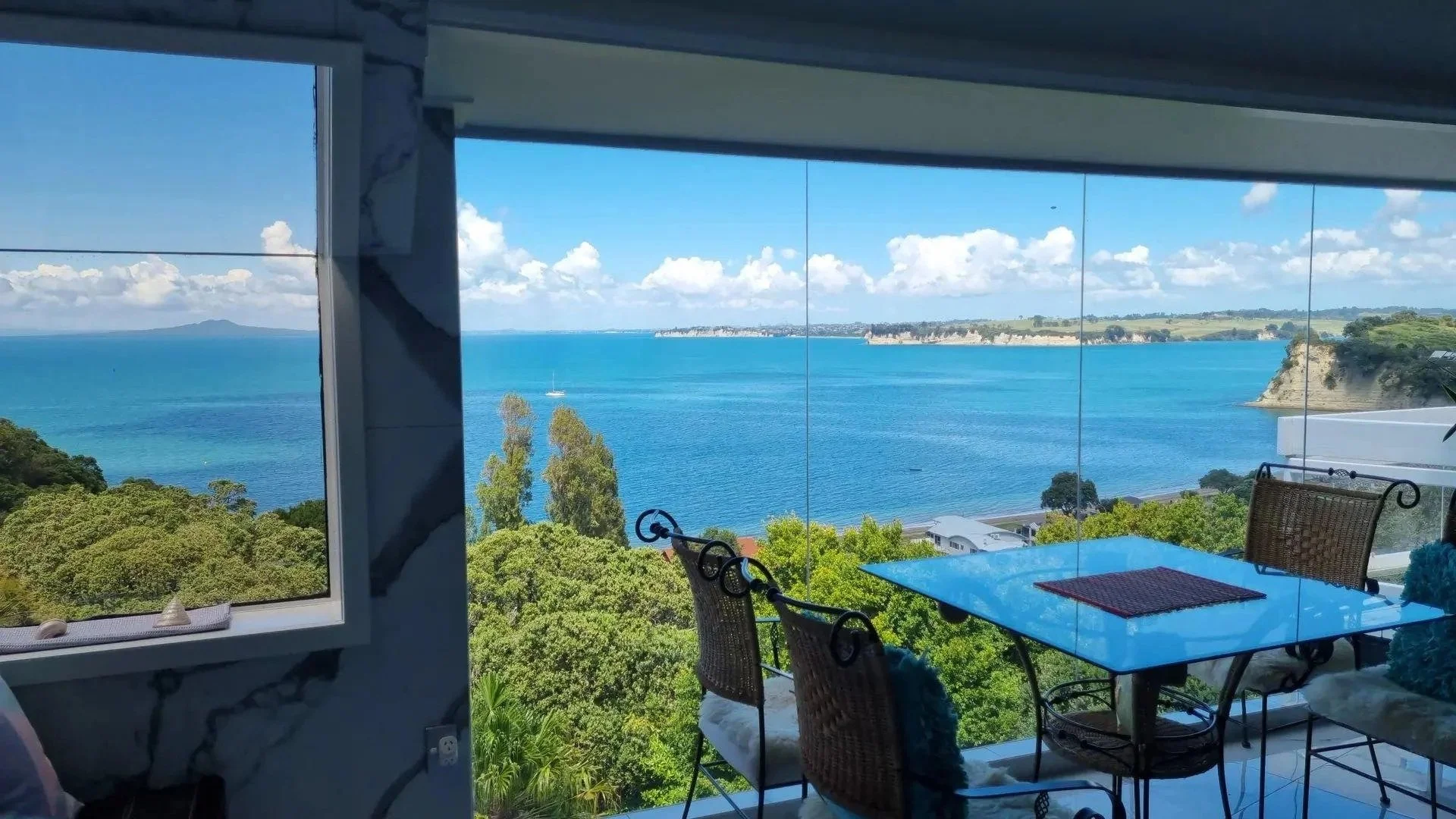 Ocean view from a balcony, with trees in the foreground, blue water, sailboats, and distant cliffs under a partly cloudy sky.