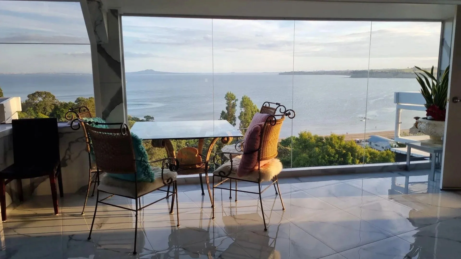 Room with a large window view of a lake, trees, boats, and distant land; interior includes a glass-top table, four chairs with cushions, and potted plants.