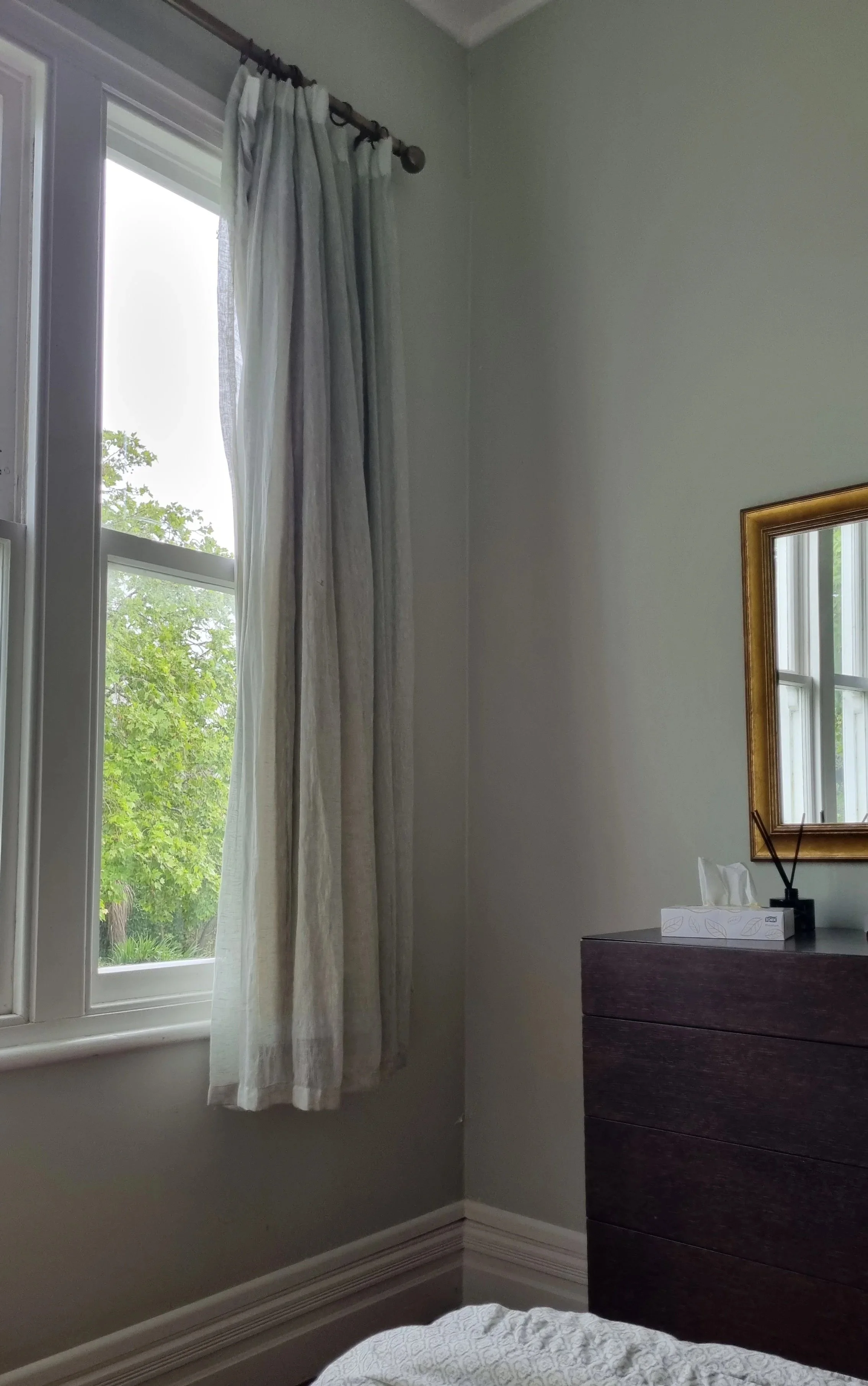 A bedroom corner with a window covered by light-colored curtains, a dresser with a tissue box and a decorative object, and part of a bed with a patterned blanket.