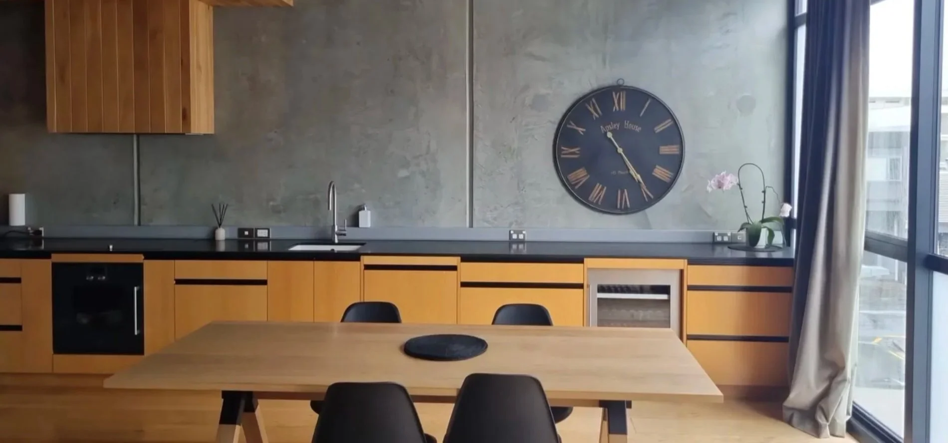Modern kitchen with wooden cabinets, a black countertop, and a round wall clock. There is a wooden dining table with black chairs, a gray curtain, and a large window showing a residential building outside.