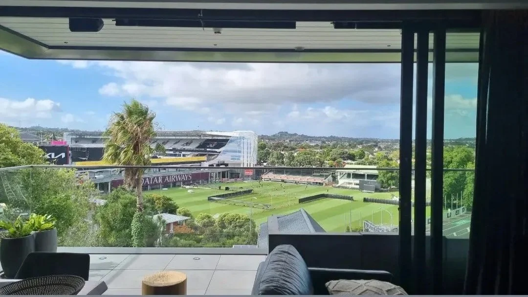 View of a cricket field from a balcony with glass railing, adjacent to a large stadium with Qatar Airways signage, under a partly cloudy sky.