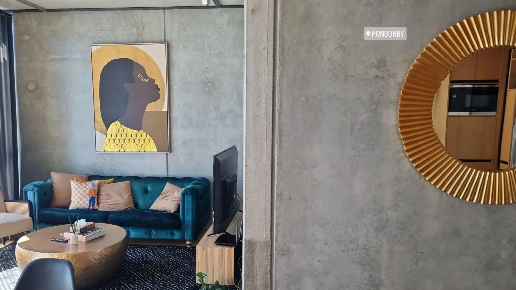 A modern living room with a teal velvet sofa, beige and patterned throw pillows, a round gold textured coffee table, a flat-screen TV on a wooden stand, an abstract portrait of a woman on the wall, and a large round gold-framed mirror on the wall.