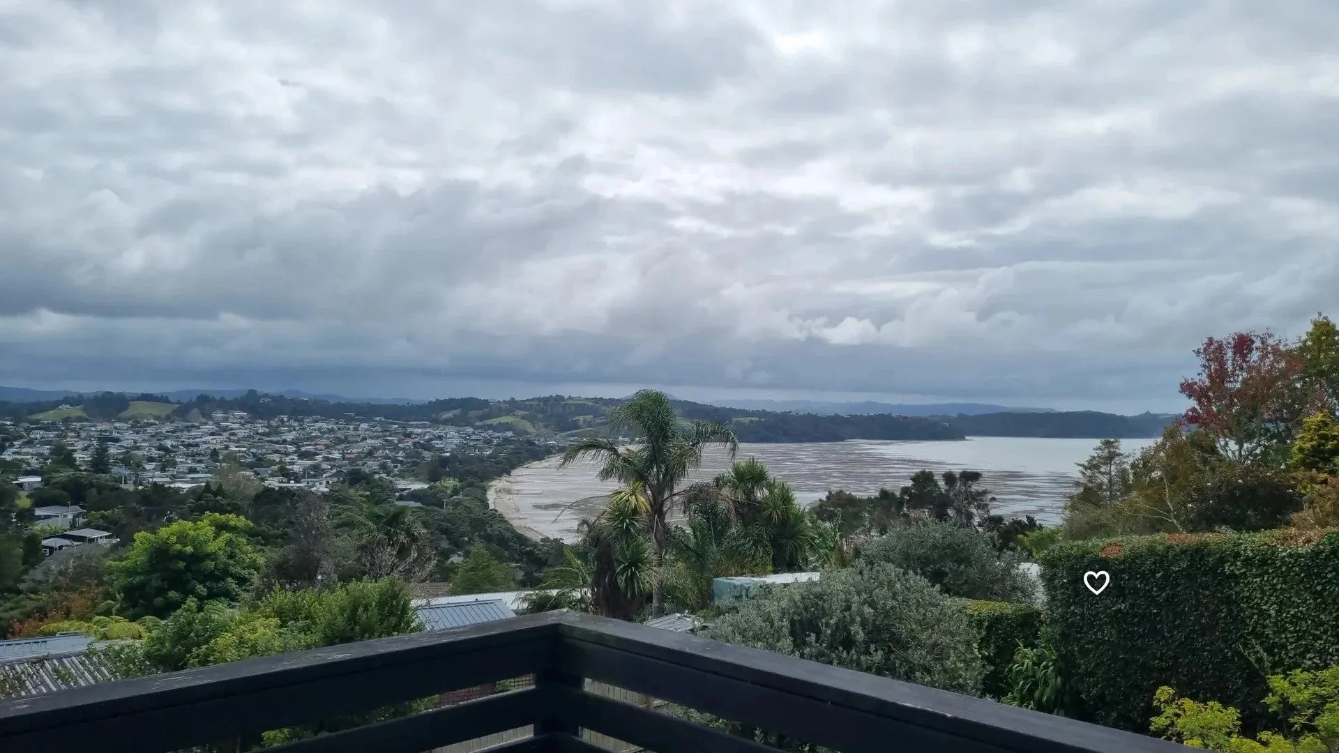 View of a cloudy sky over a suburban area with houses, trees, and a body of water in the distance.