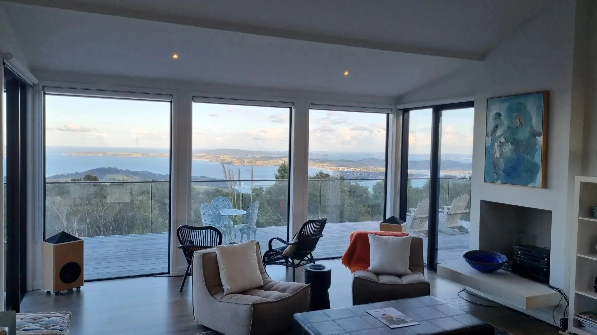 Living room with large windows overlooking a body of water and mountains, furnished with chairs, a sofa, and a colorful abstract painting on the wall.