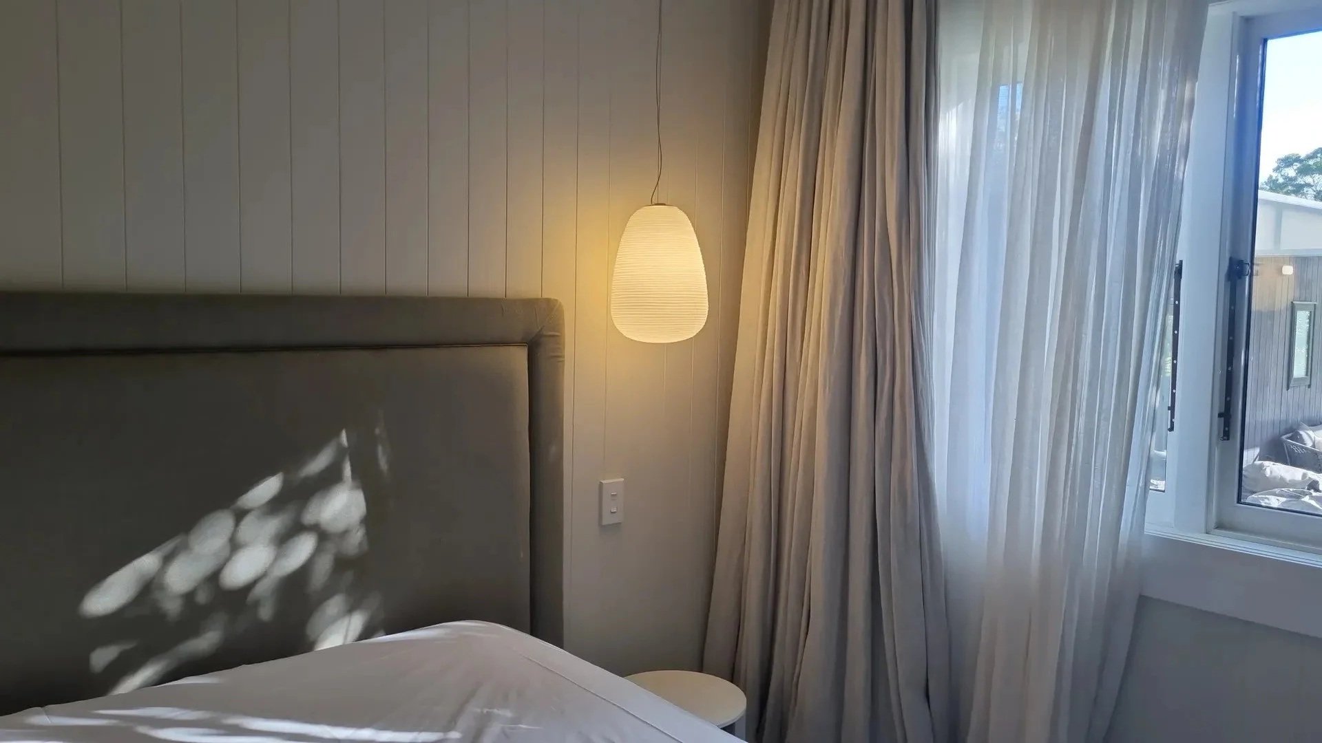 Part of a bedroom showing a bed, a beige headboard, a hanging paper lantern, a white curtain, and an open window with a view outside.