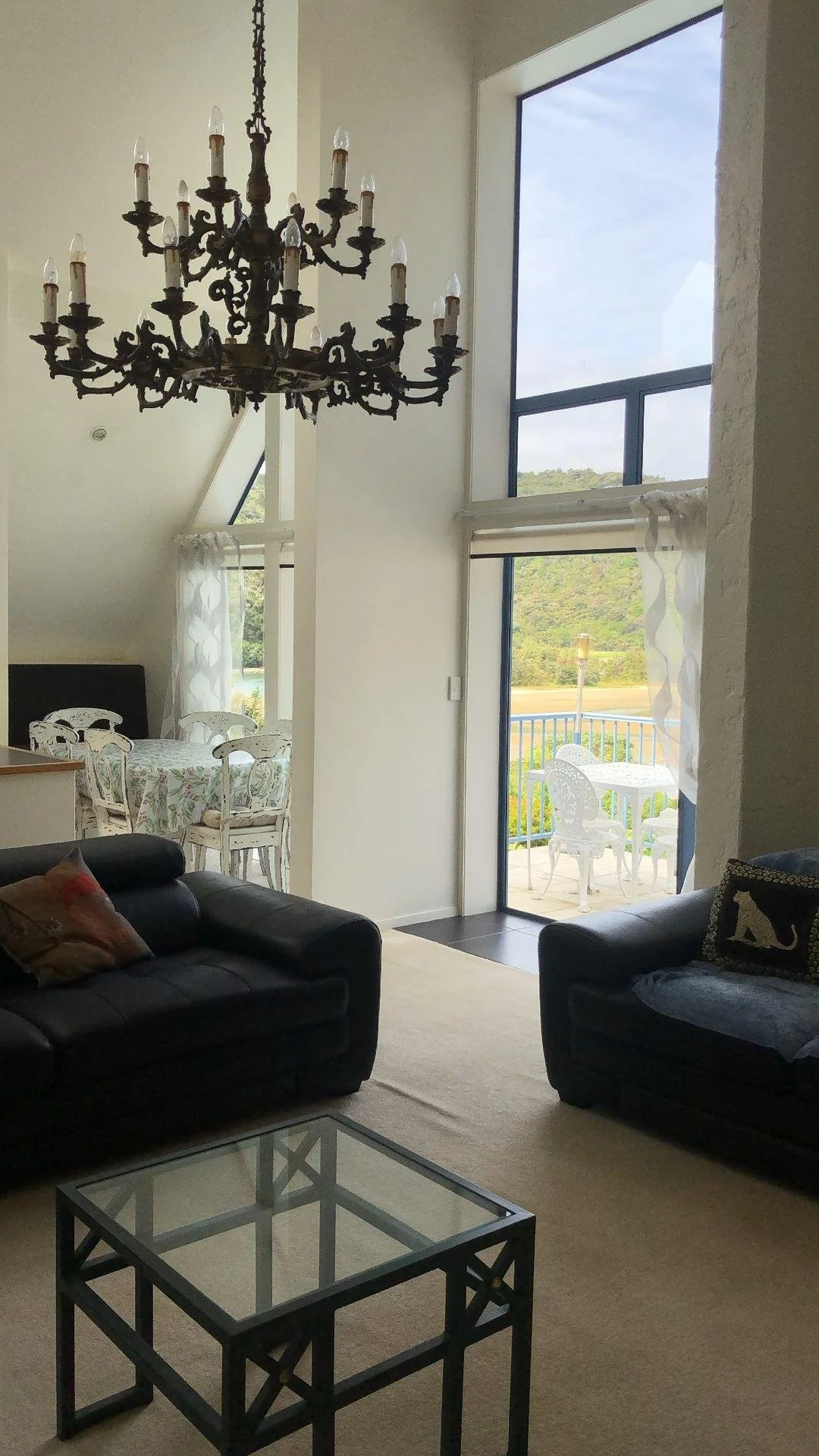 Living room with dark leather couches, glass coffee table, chandelier, large window with curtains, and a dining area with a round table and chairs outside on a balcony.
