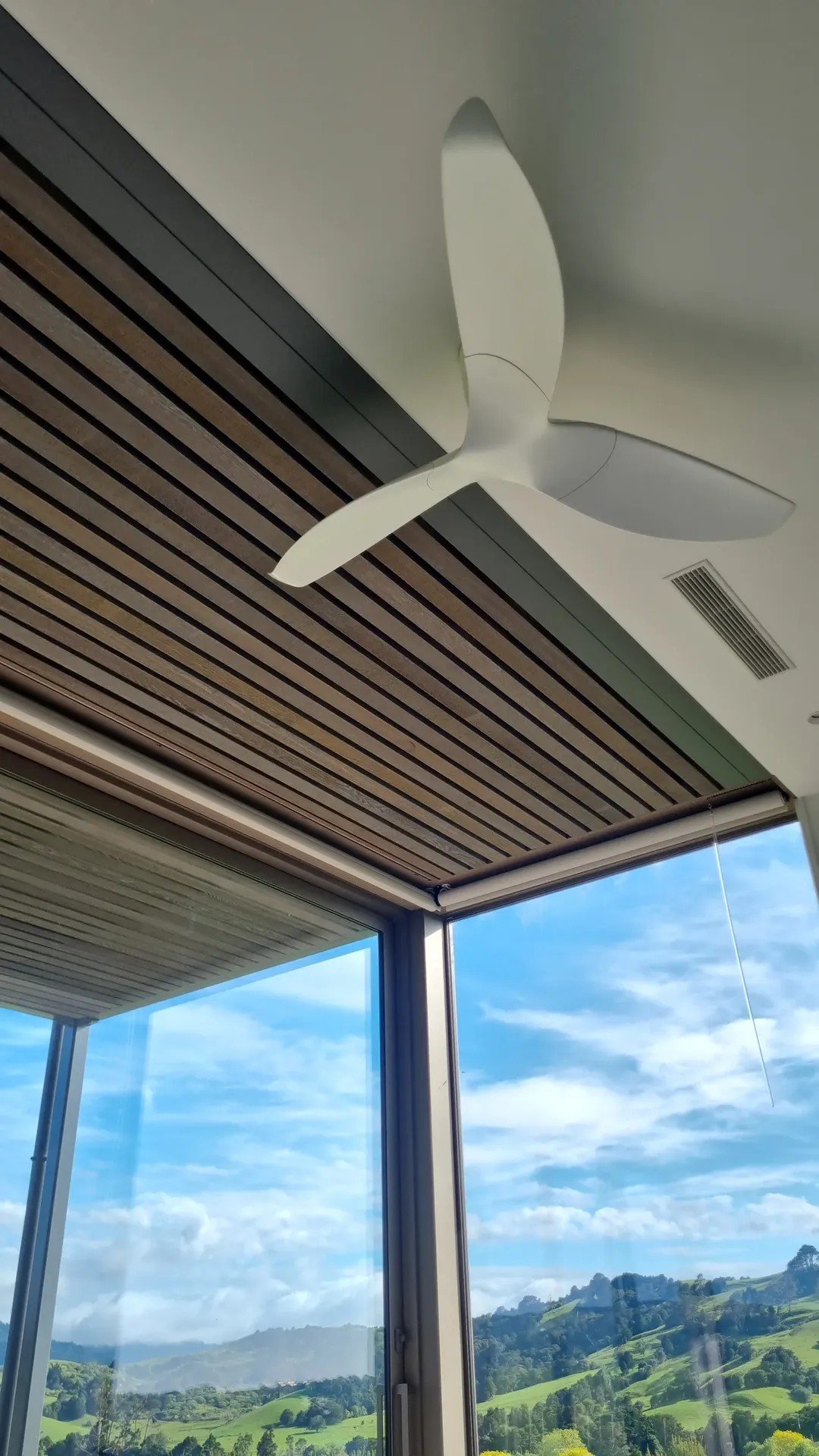 Interior view of a room with a white ceiling fan, wooden slatted ceiling, and large windows showing a scenic landscape of rolling green hills and blue sky with clouds.