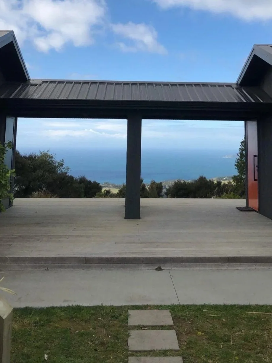 View through a modern house opening showing a distant ocean and sky with scattered clouds, framed by a metal roof and dark-colored walls.